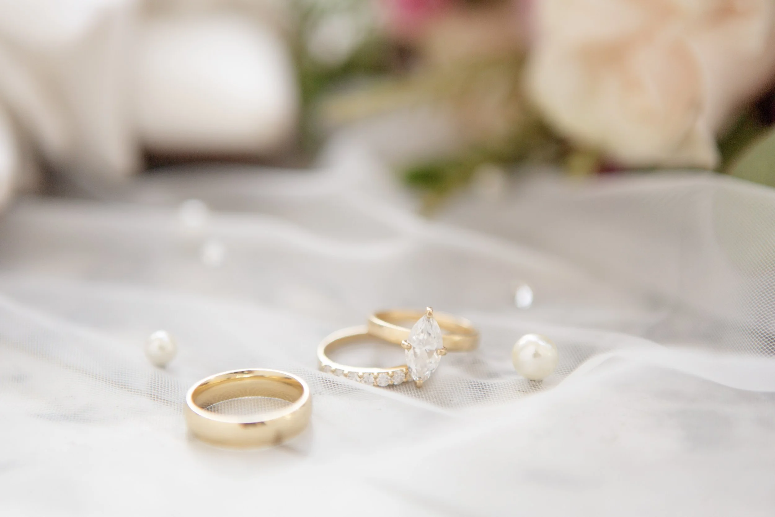 Close-up of a wedding ring and engagement ring, a pearl, and a diamond ring, with a blurred background of white petals and greenery.