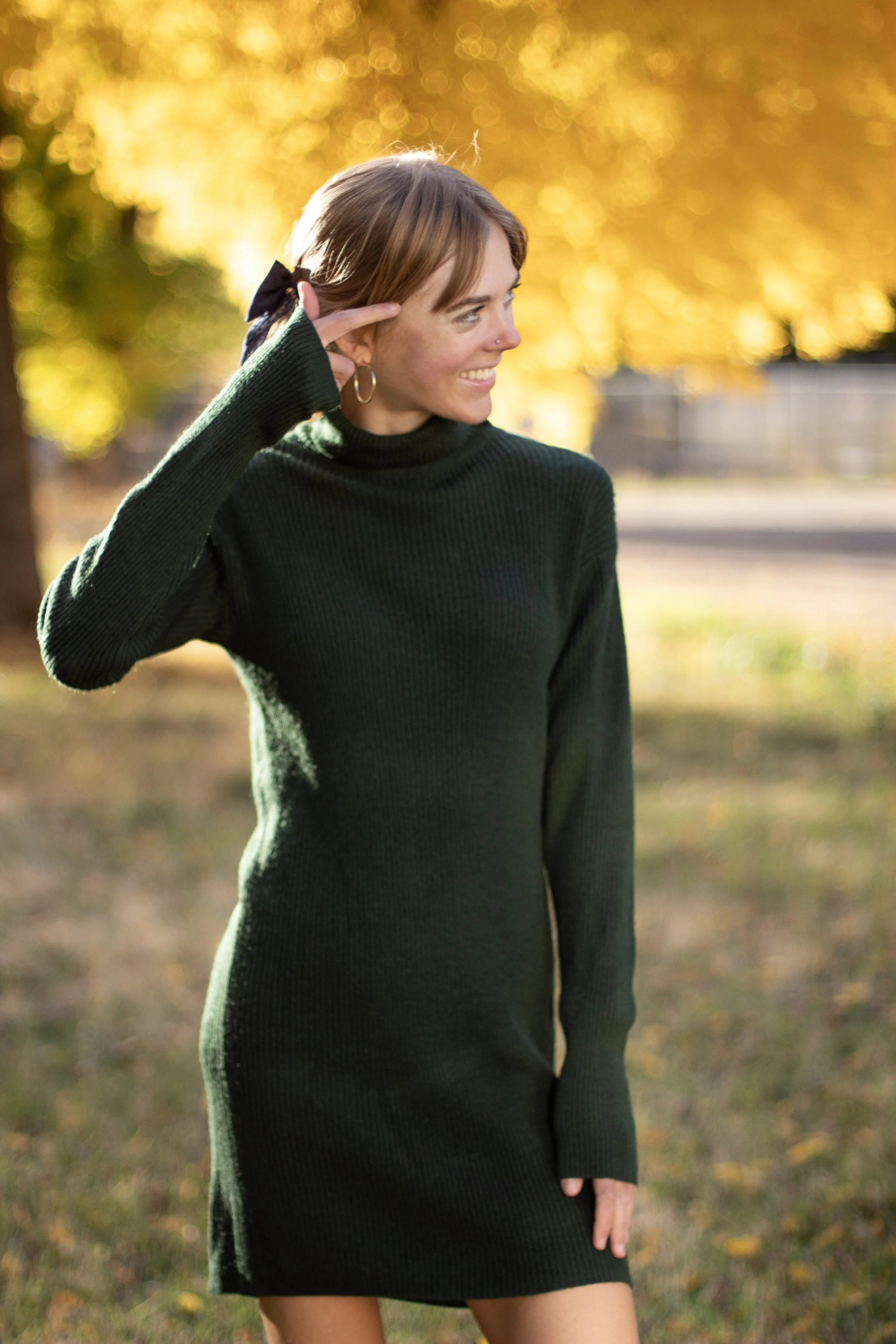 A young woman in a black sweater dress standing outdoors in a park with autumn foliage background, smiling and making a peace sign near her head.