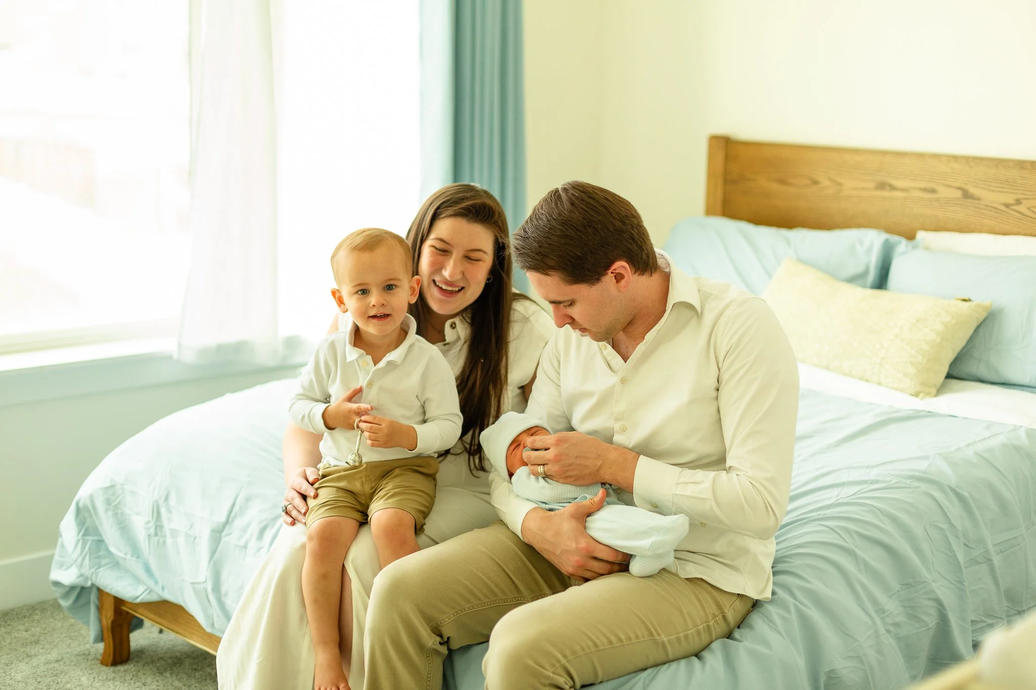 Family with parents and two young children gathered on bed in bright room, celebrating the birth of a newborn baby.