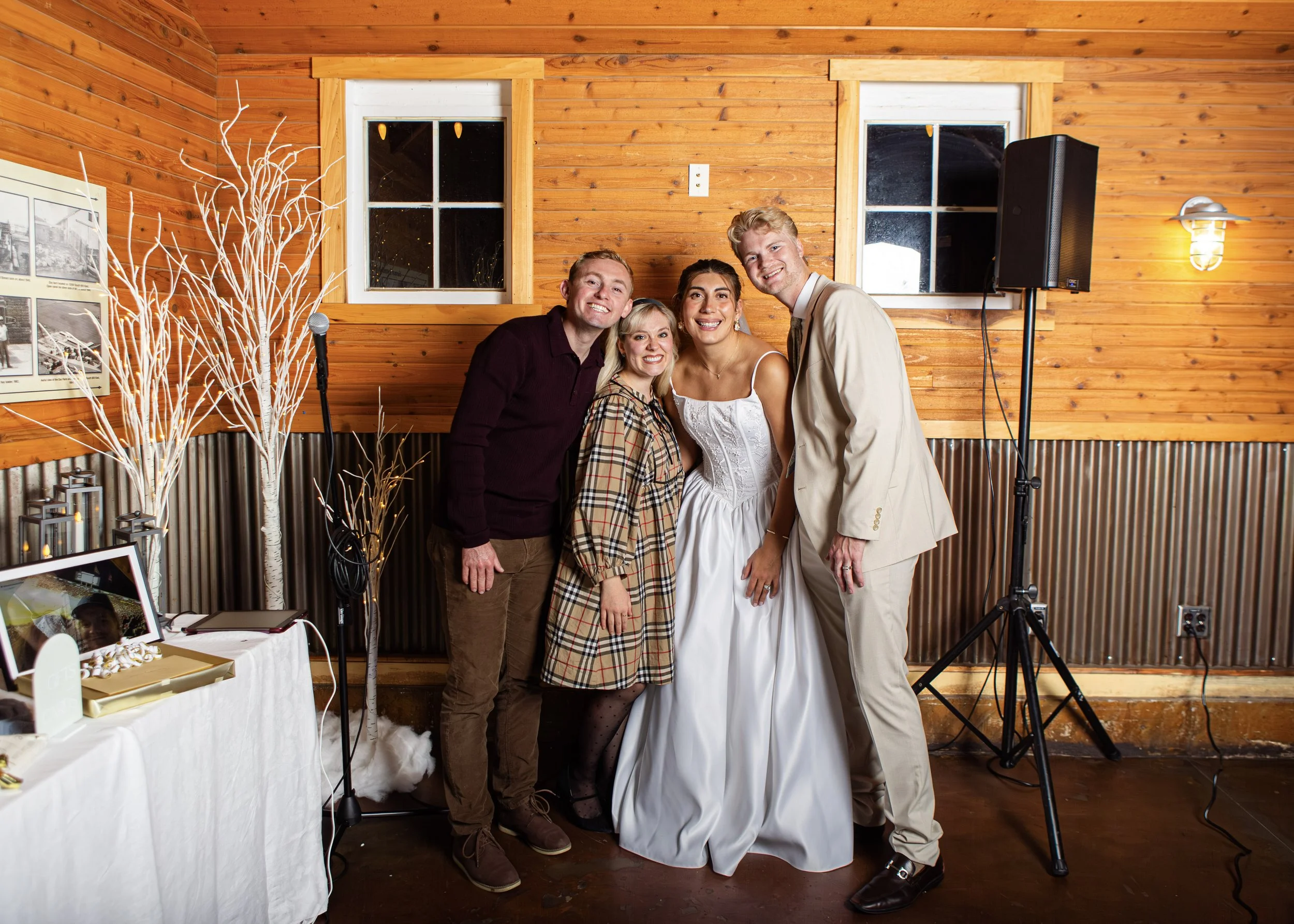 Four people smiling and posing together indoors with wooden walls, two windows, a speaker on a stand, and a decorated table with photos and gifts.