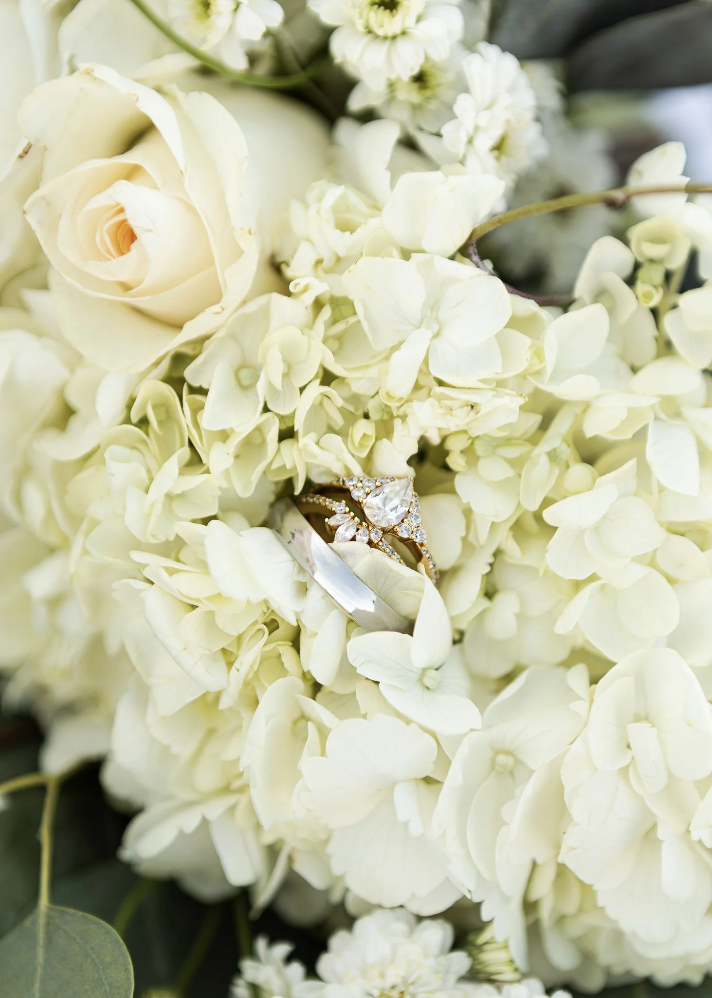 Close-up of a bouquet of white roses, hydrangeas, and other flowers with a diamond engagement ring placed on the flowers.