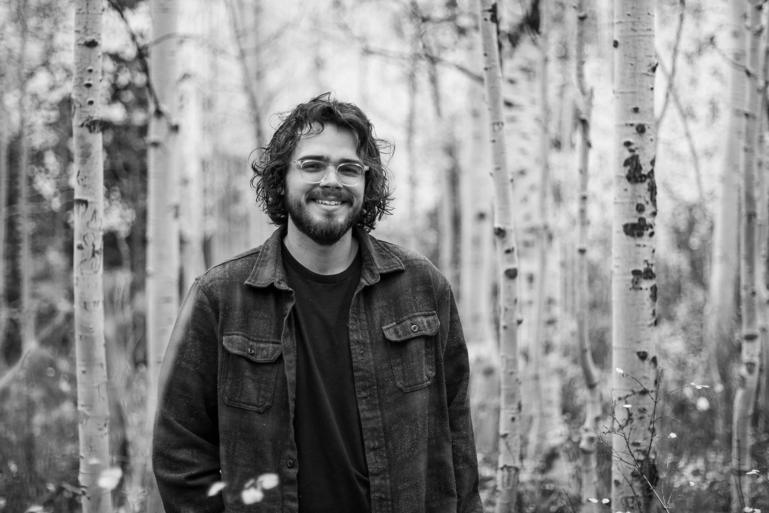 A man with curly hair, glasses, and a beard smiling while standing in a forest of aspen trees