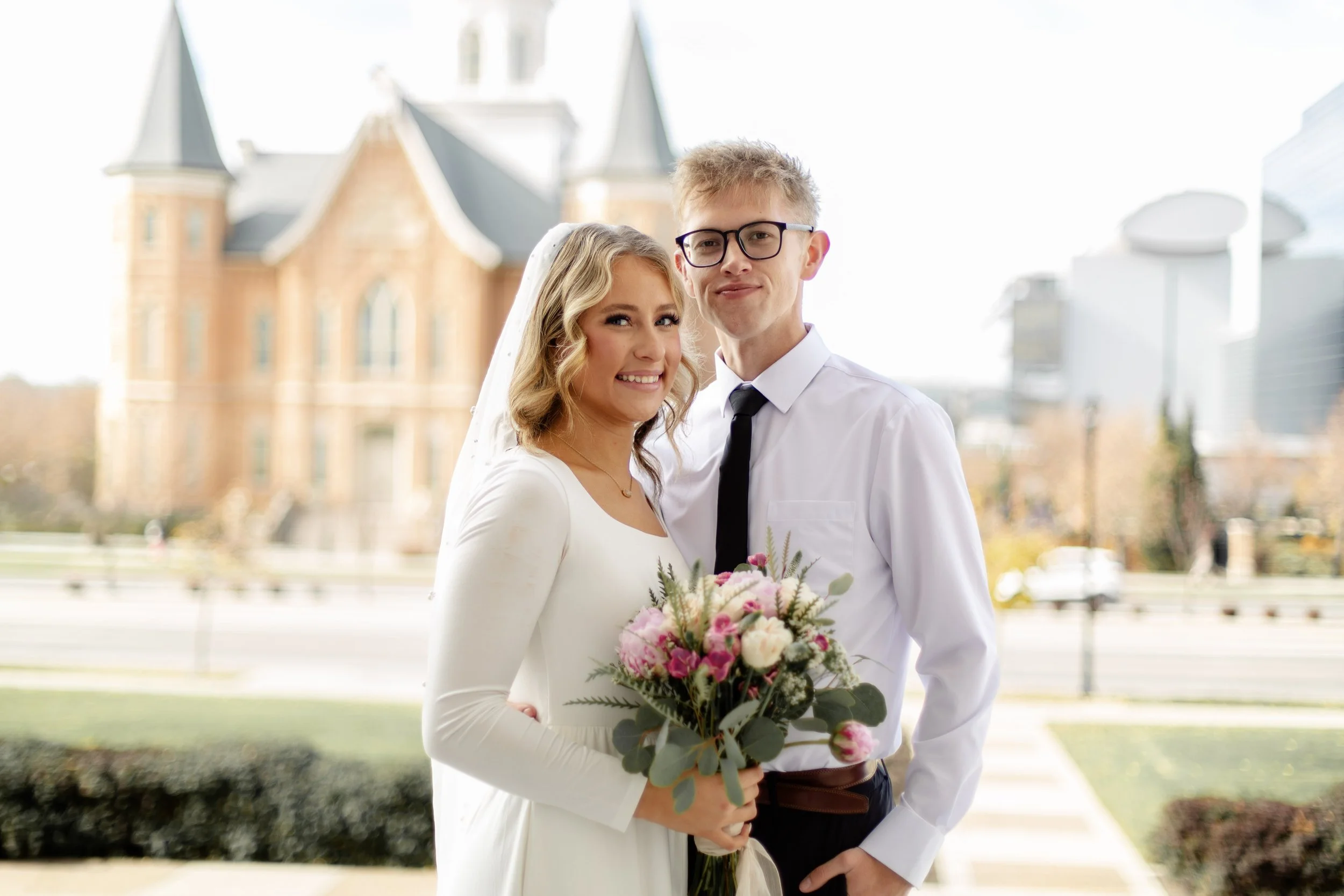 A bride and groom sharing a kiss, with the bride holding a bouquet of red roses and white baby's breath during sunset.