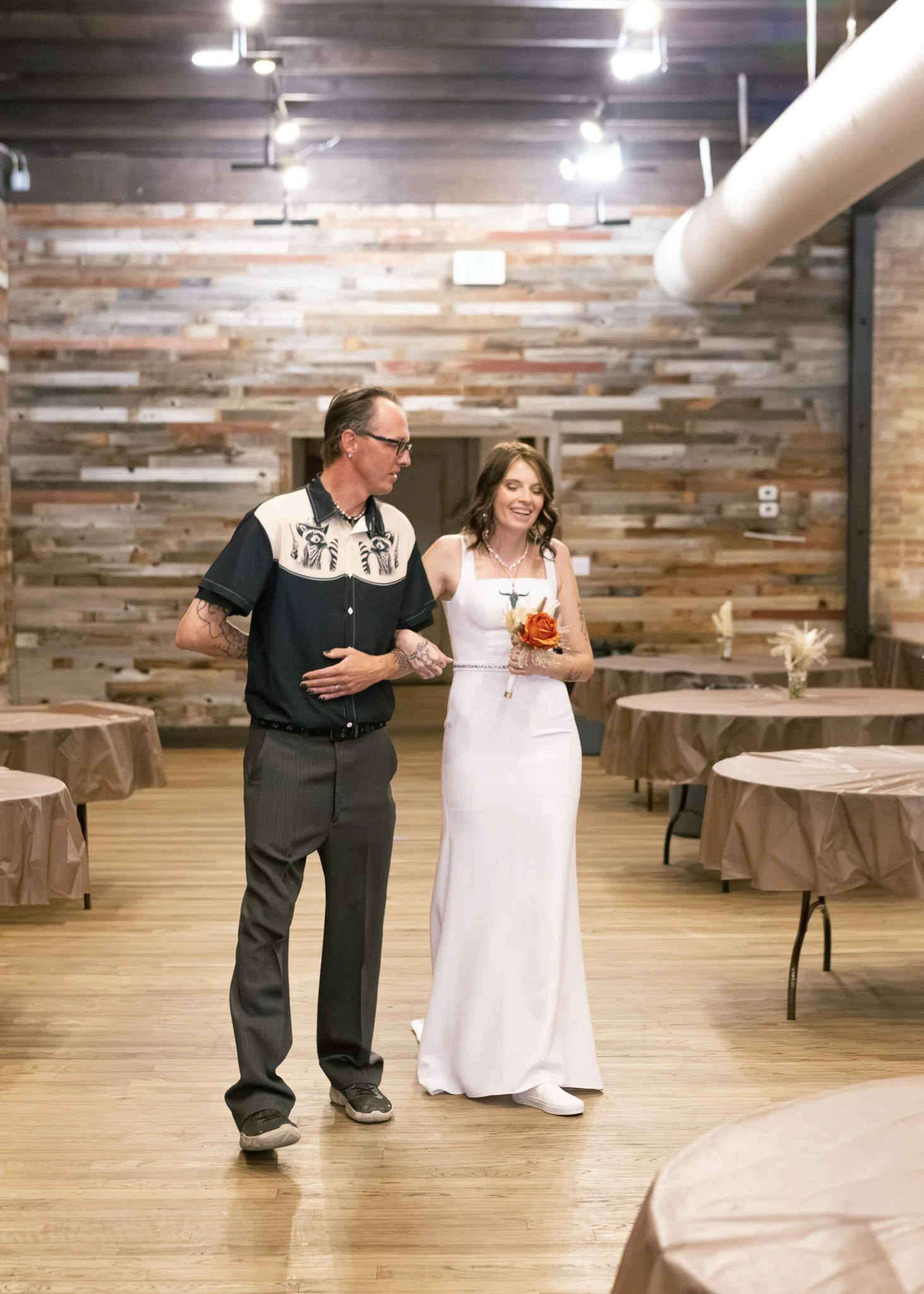 A man and woman are standing inside a rustic wedding reception venue with wooden walls and tables covered with brown tablecloths. The woman is smiling and holding a bouquet, wearing a white dress, and the man has tattoos on his arms and is wearing gl