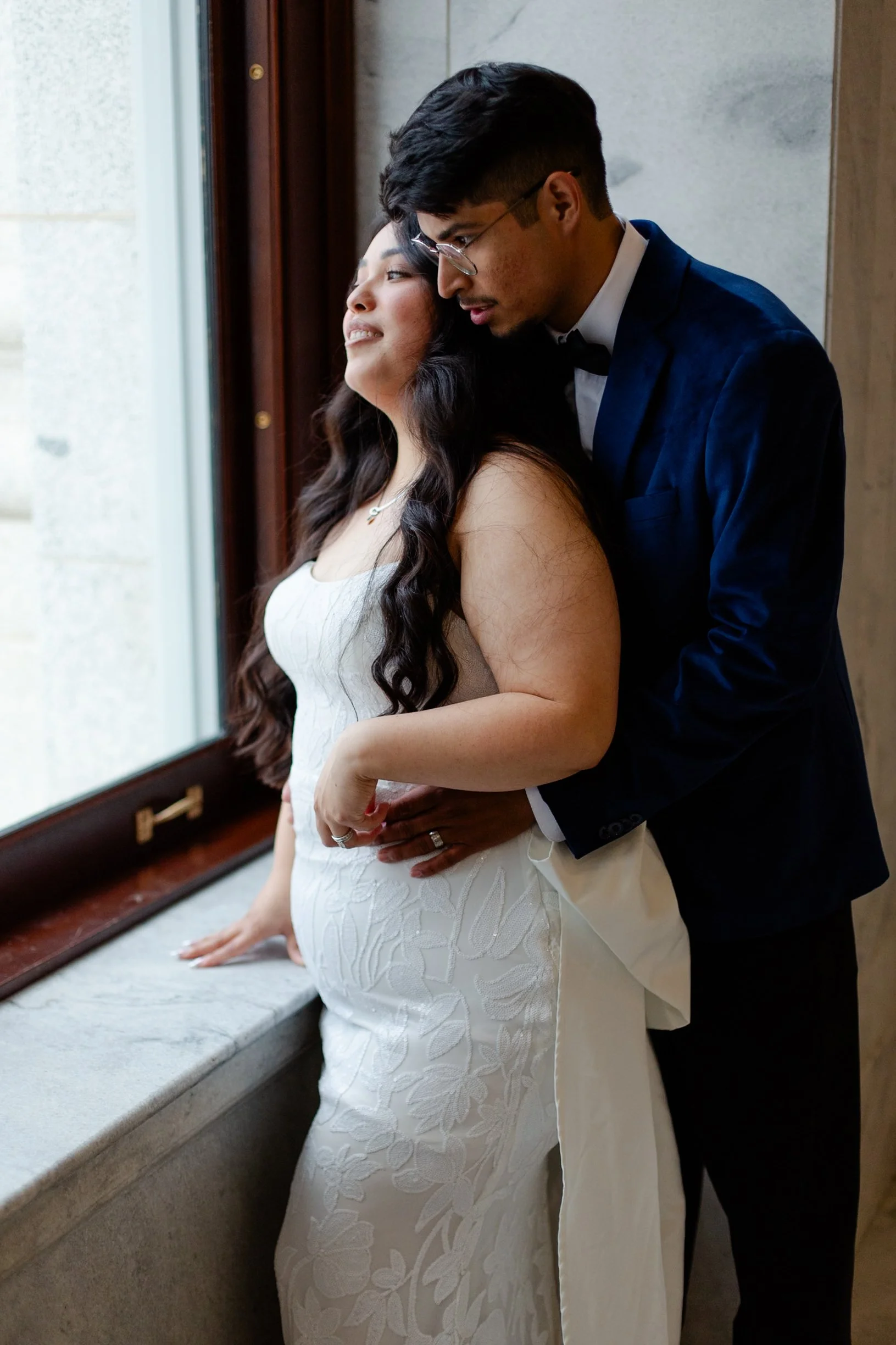 A newlywed couple standing by a window, with the bride in a white lace wedding dress and the groom in a dark suit with a bow tie, embracing each other as they look out the window.