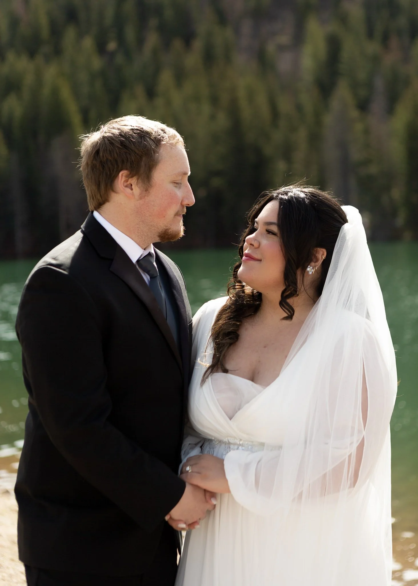 A groom and bride stand close together outdoors, holding hands and smiling at each other. The groom wears a black suit with a white shirt and black tie. The bride wears a white wedding dress with a veil and floral earrings. They are by a lake with a 