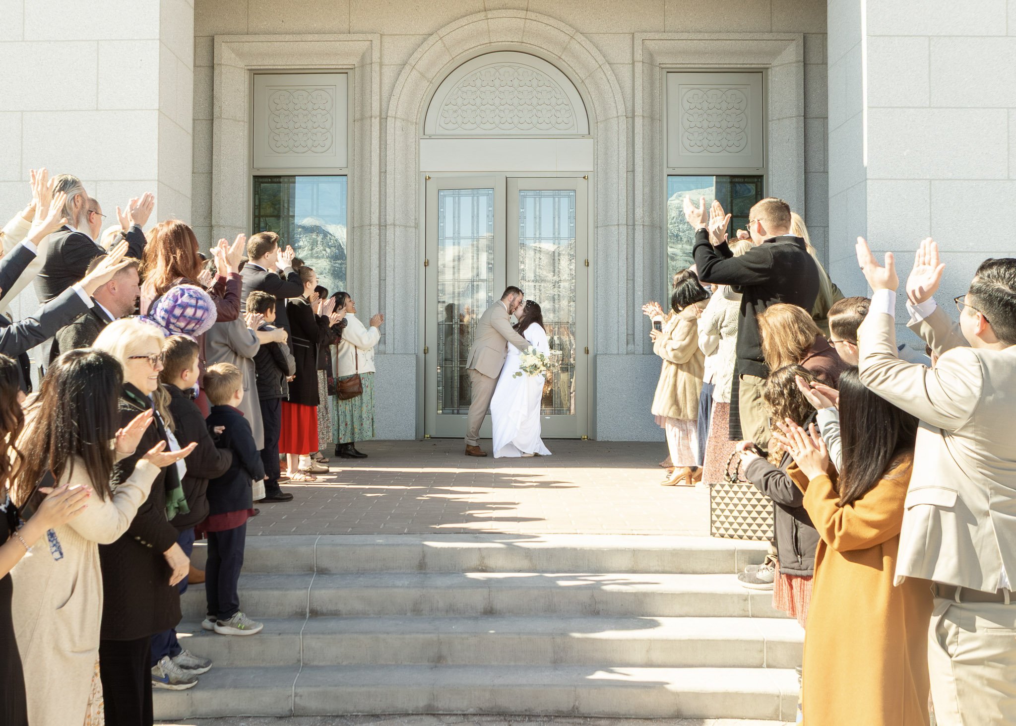 A bride and groom kissing under a glass door. Guests on both sides of the stairs are clapping and celebrating.