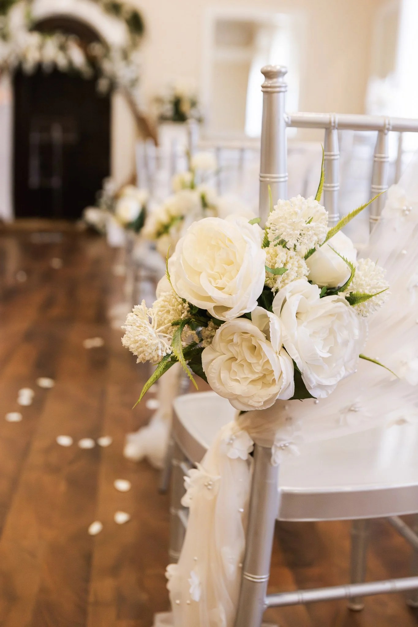 Close-up of a chair with white floral decorations, set in a decorated wedding or event venue with scattered white petals on the wooden floor.