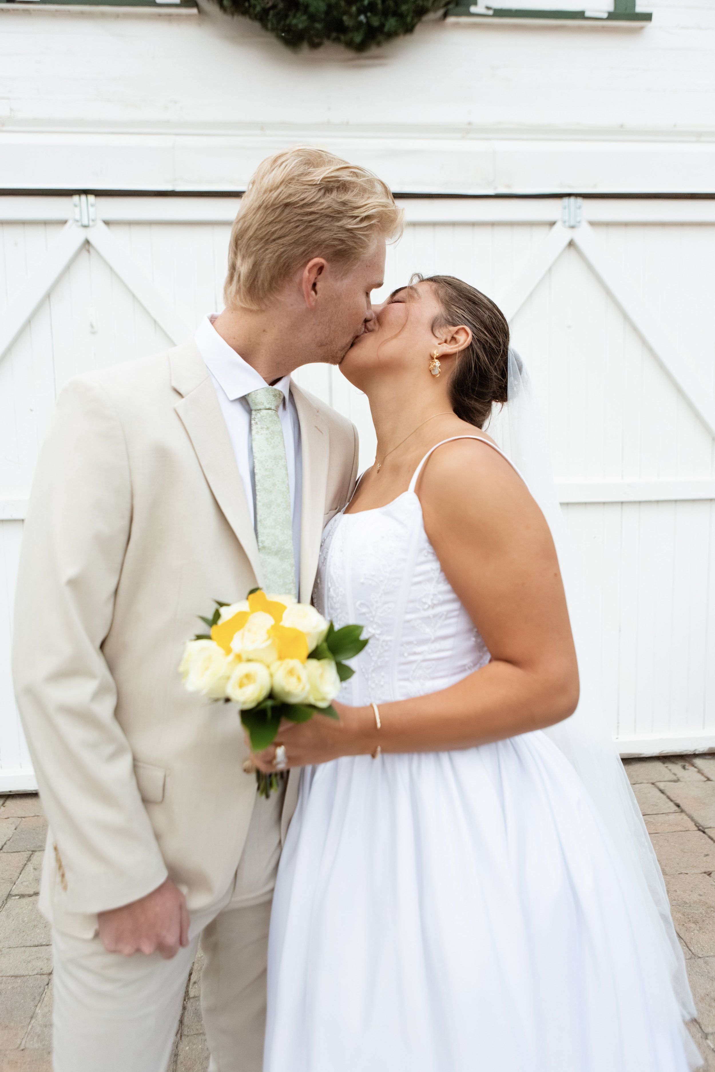 A newlywed couple sharing a kiss, with the groom holding a bouquet of yellow and white roses, standing in front of a white barn door.