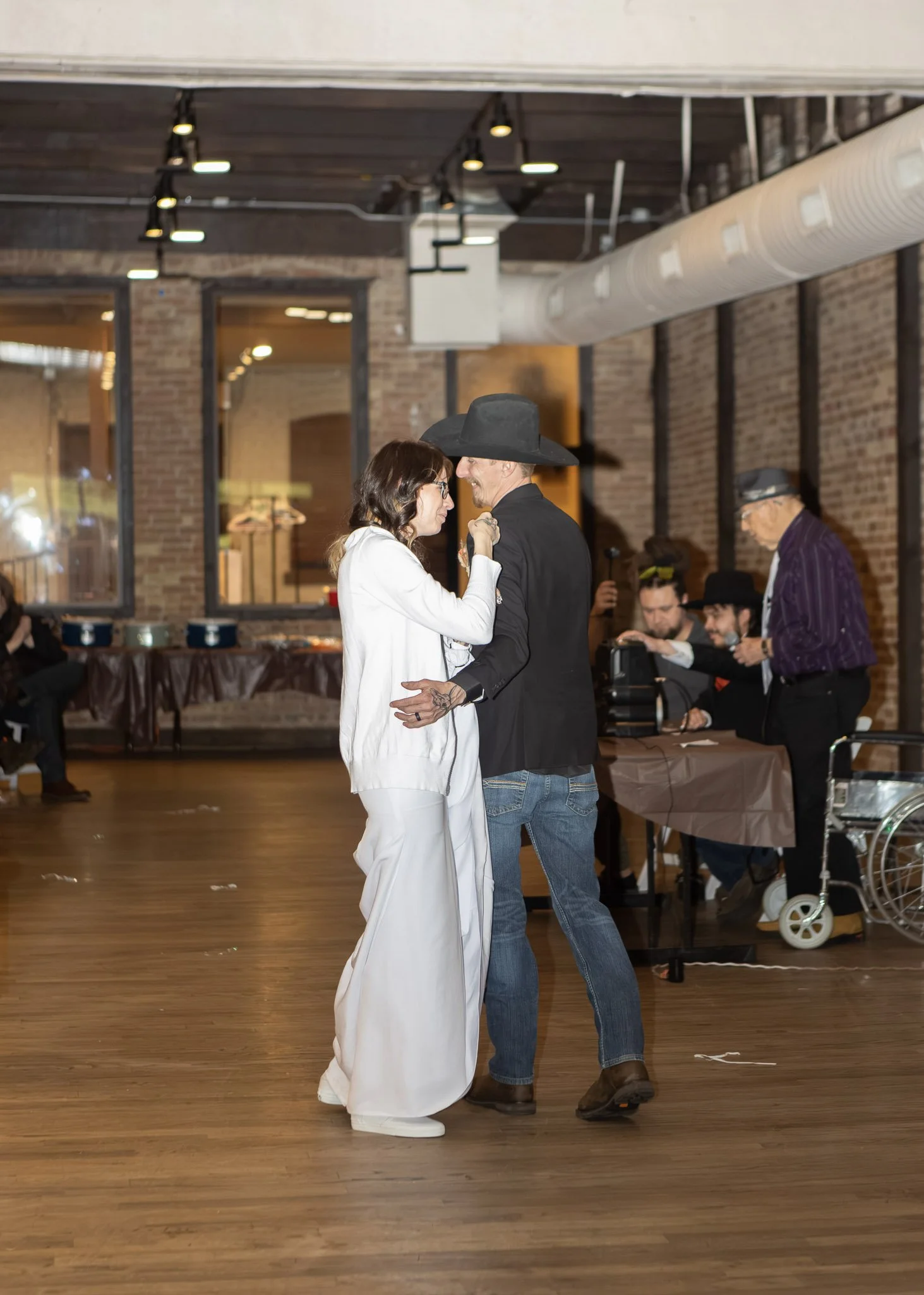 A couple dancing indoors with brick walls and wooden floors, the woman wearing a white outfit and glasses, the man in a black shirt and large black hat, surrounded by people including an elderly person with glasses and a purple striped shirt in the b
