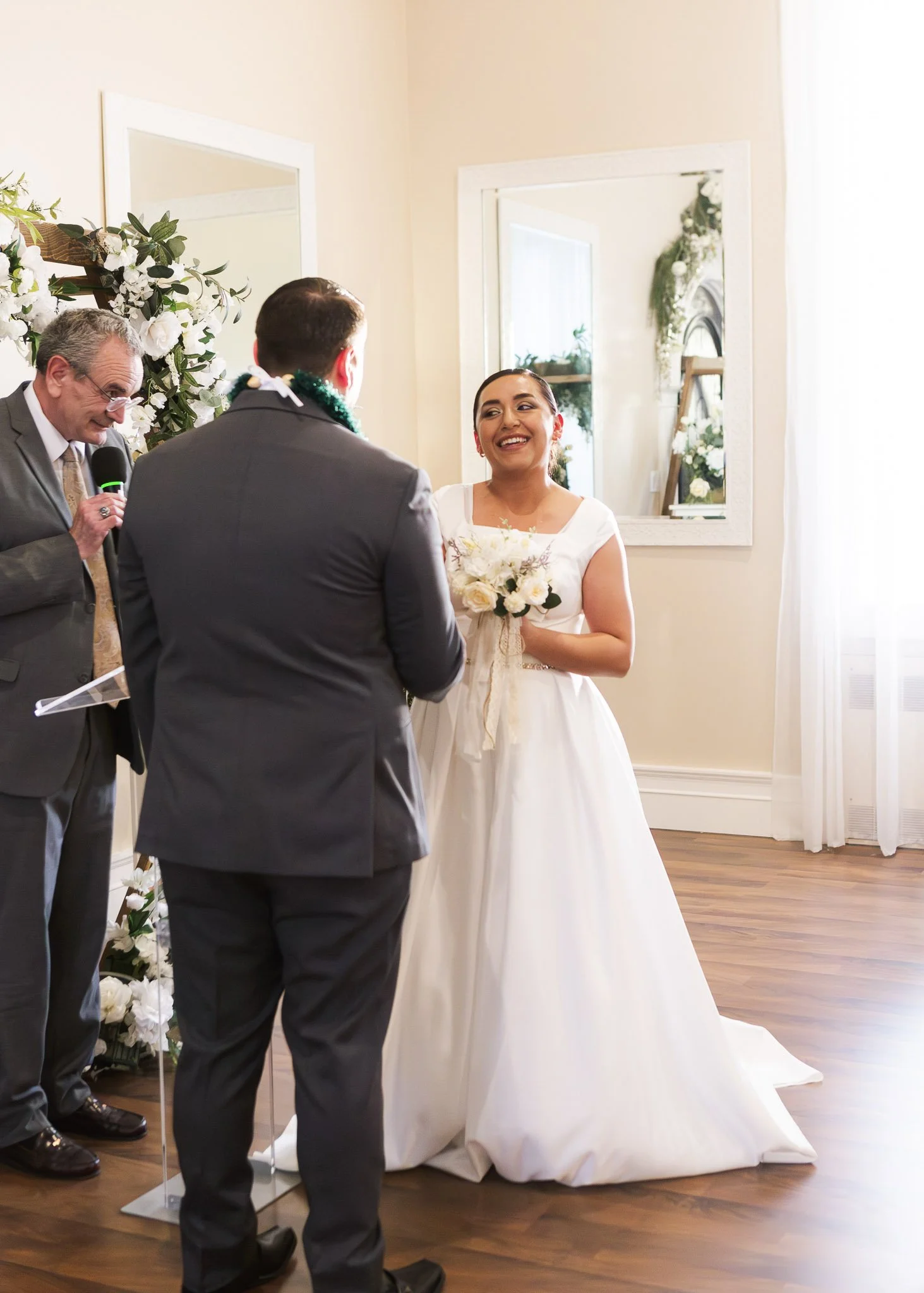 A bride in a white wedding dress holding a bouquet, smiling during her wedding ceremony with two men, one with gray hair and glasses holding a microphone, and another with dark hair seen from the back, all in an indoor setting with large mirrors and 