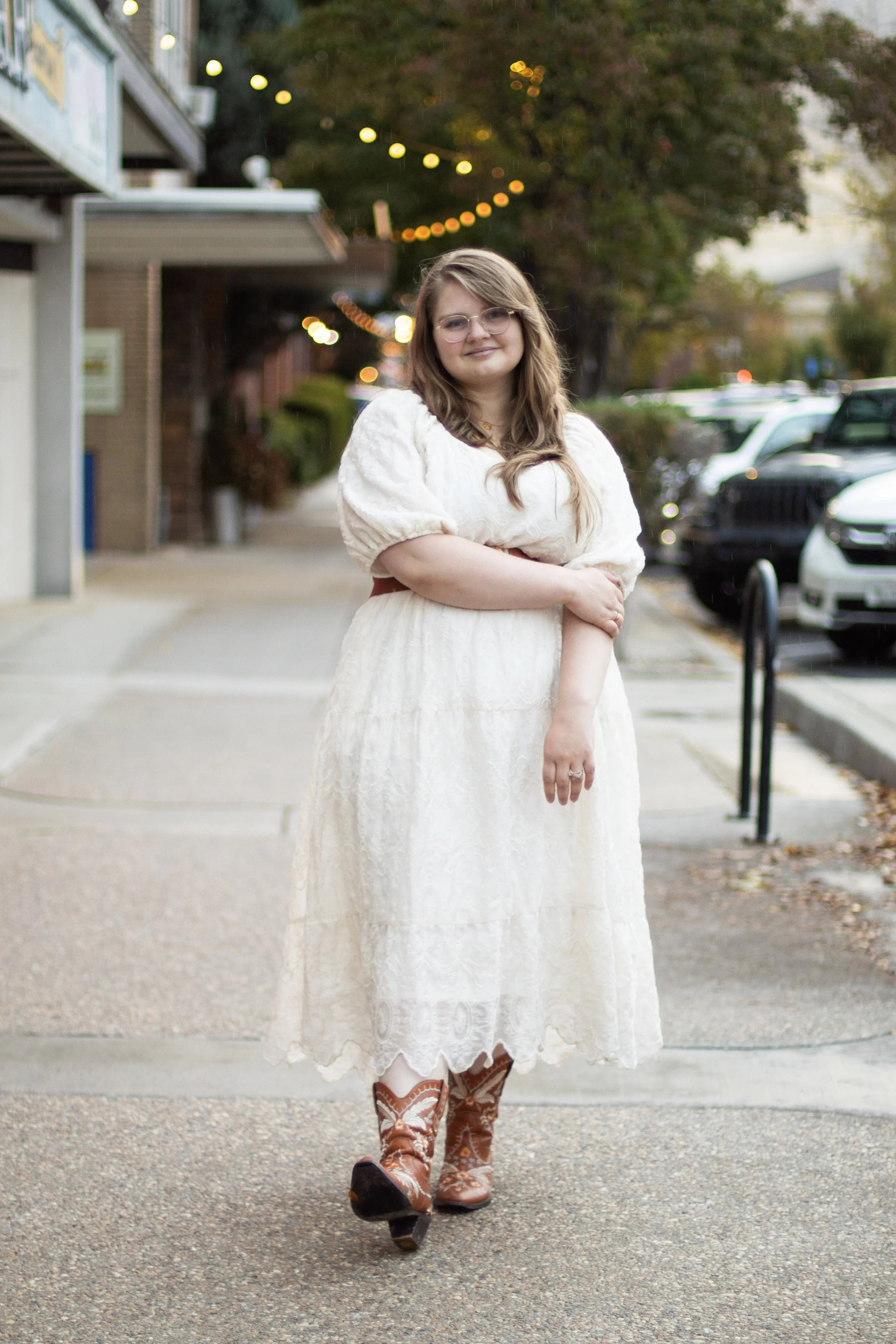 Young woman standing on sidewalk in autumn, wearing a vintage white lace dress and cowboy boots, with blurred string lights and parked cars in the background.