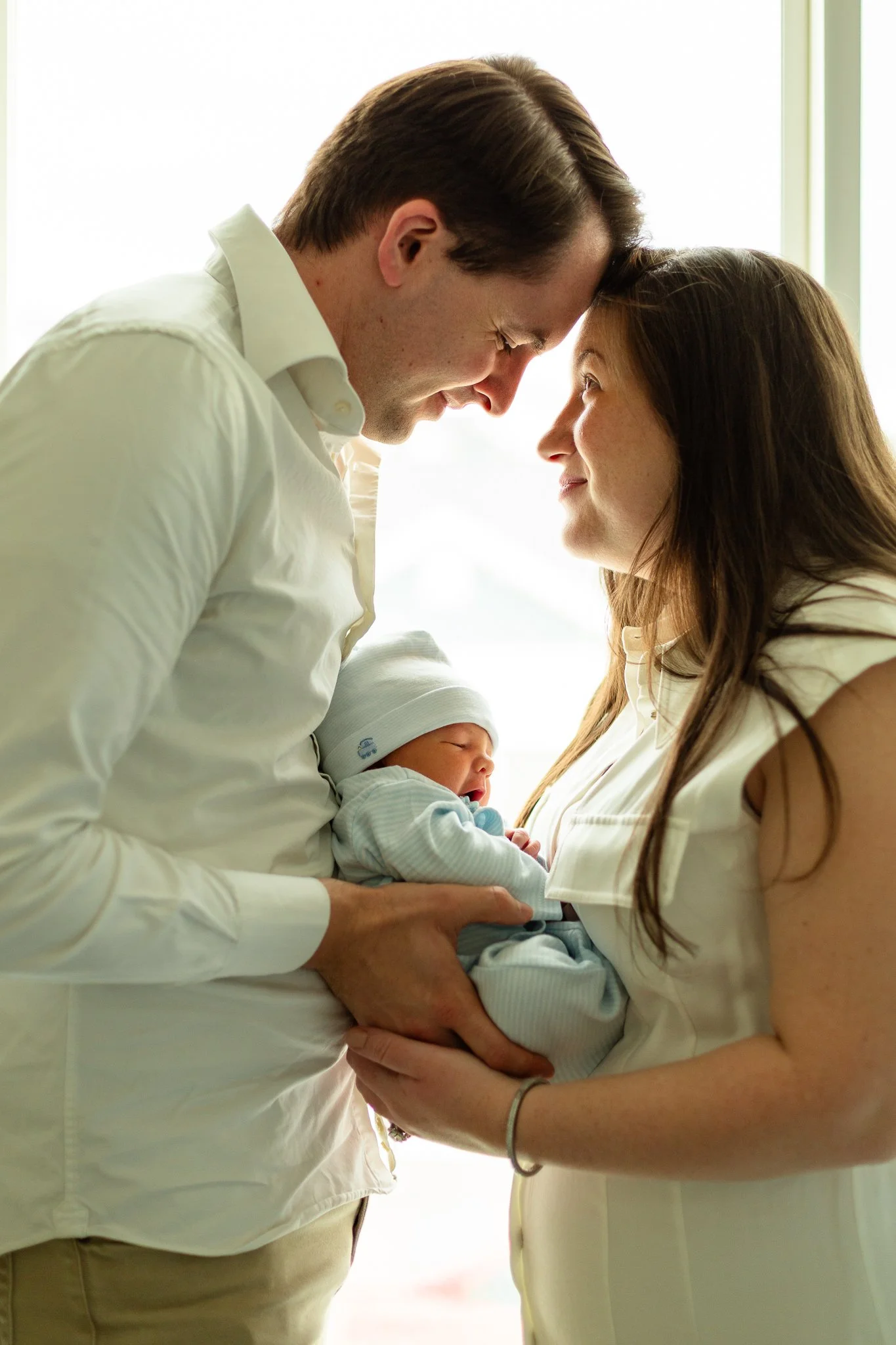 A happy couple holding their newborn baby close to their foreheads, with the mother holding the baby in her arms, in front of a bright window.
