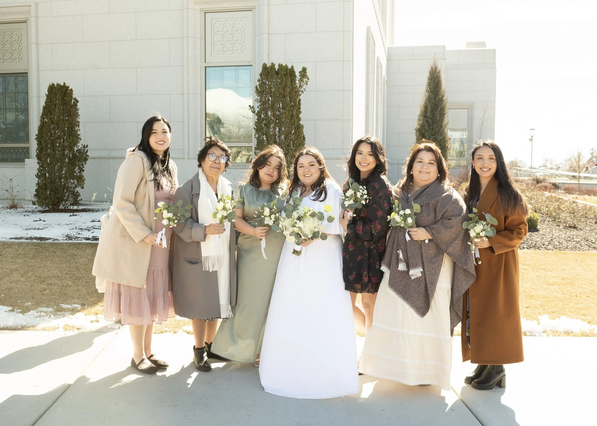 Group of women standing outdoors, dressed in formal and semi-formal attire, holding bouquets of white flowers, with a modern building and trees in the background on a clear, sunny day.