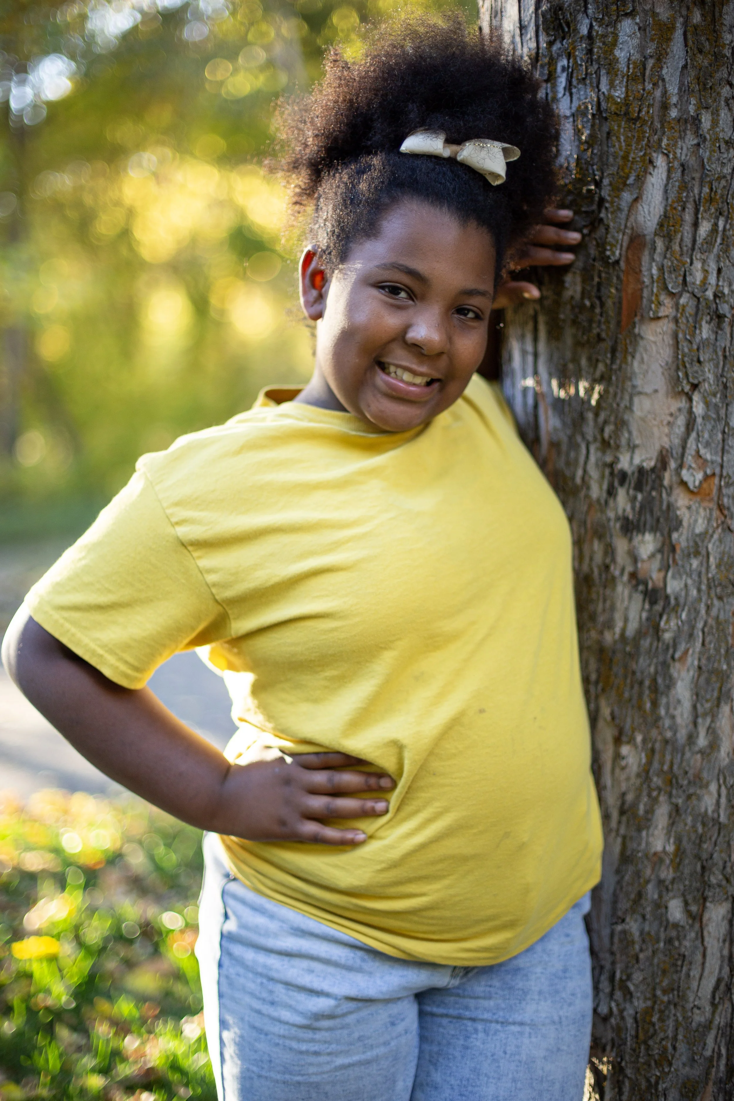 Young girl wearing a yellow T-shirt and light blue jeans smiling, standing outdoors next to a tree with sunlight filtering through the leaves.