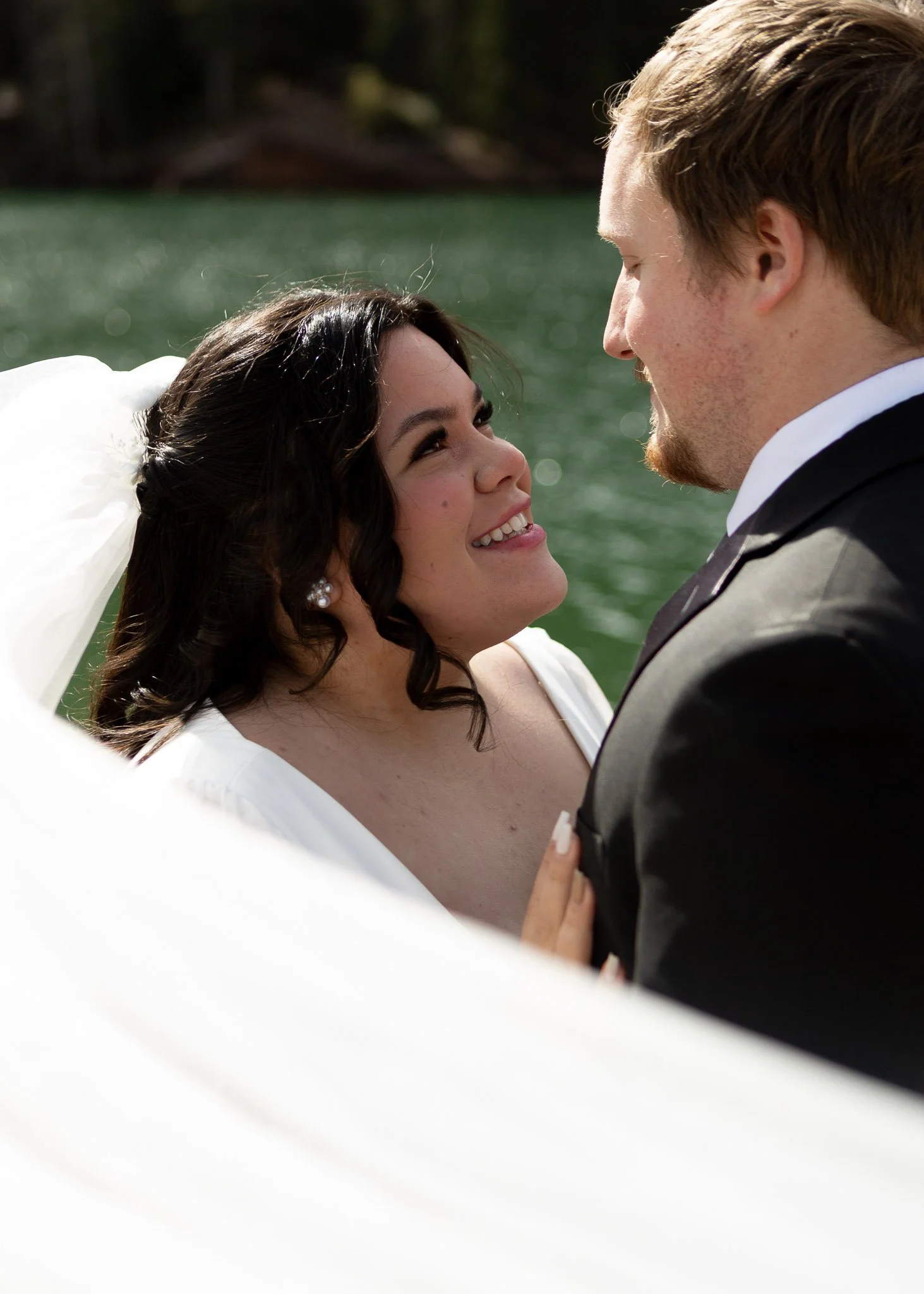 A bride and groom gazing at each other on their wedding day, outdoors near a body of water with trees in the background.