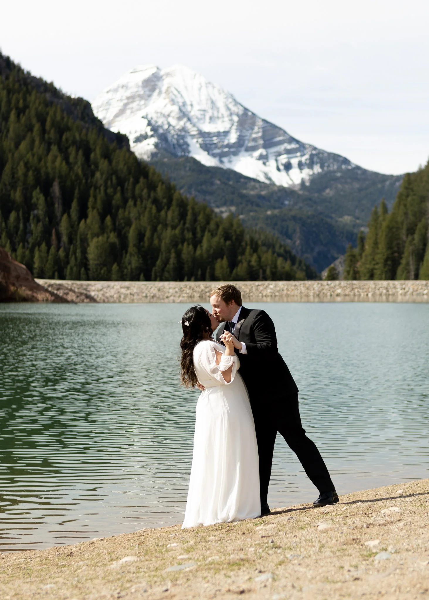 A bride and groom standing by a lake with snow-capped mountain in the background, sharing a kiss.