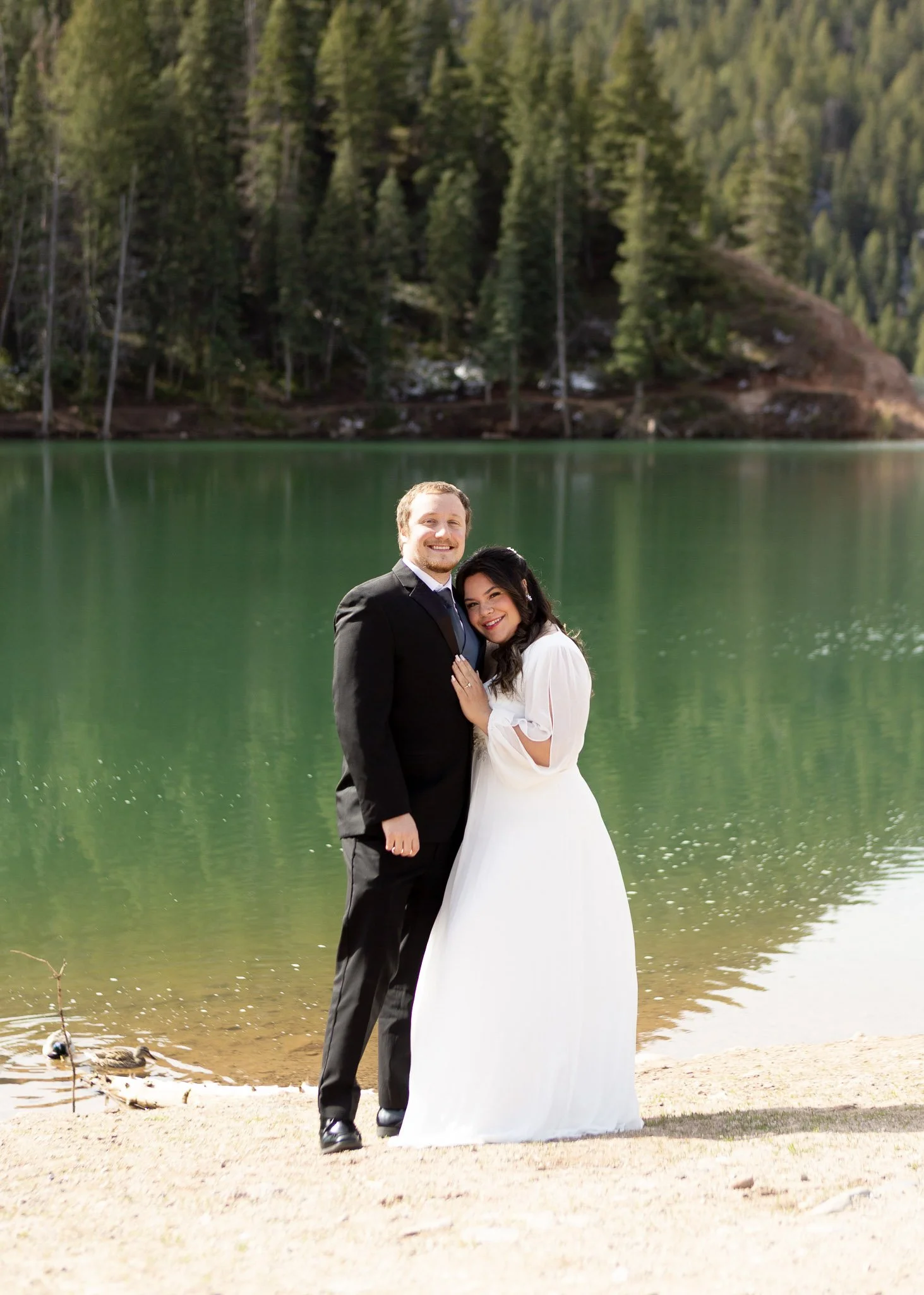 A bride and groom standing together by a lake in a forested area, smiling at the camera.