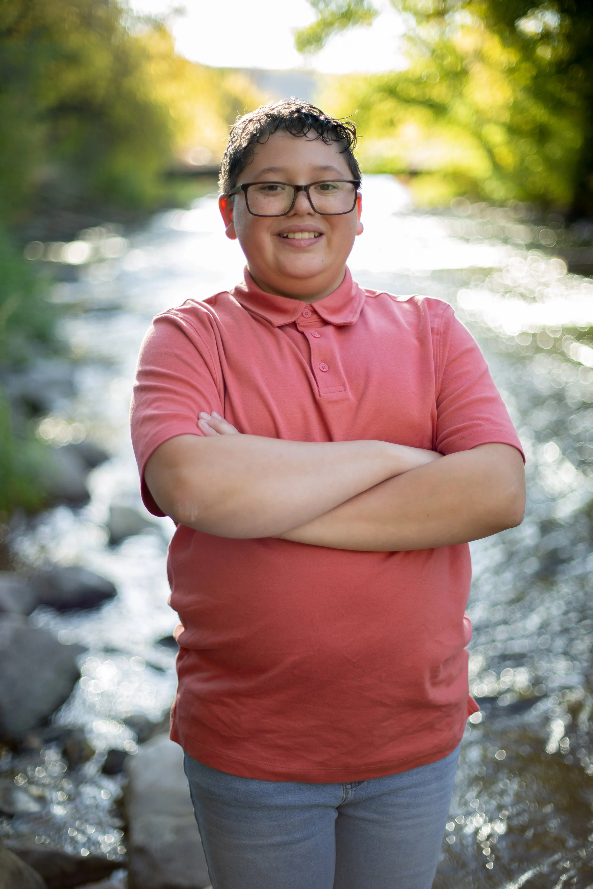 A young boy with glasses and a pink polo shirt standing with arms crossed outdoors near a creek with trees and sunlight in the background.