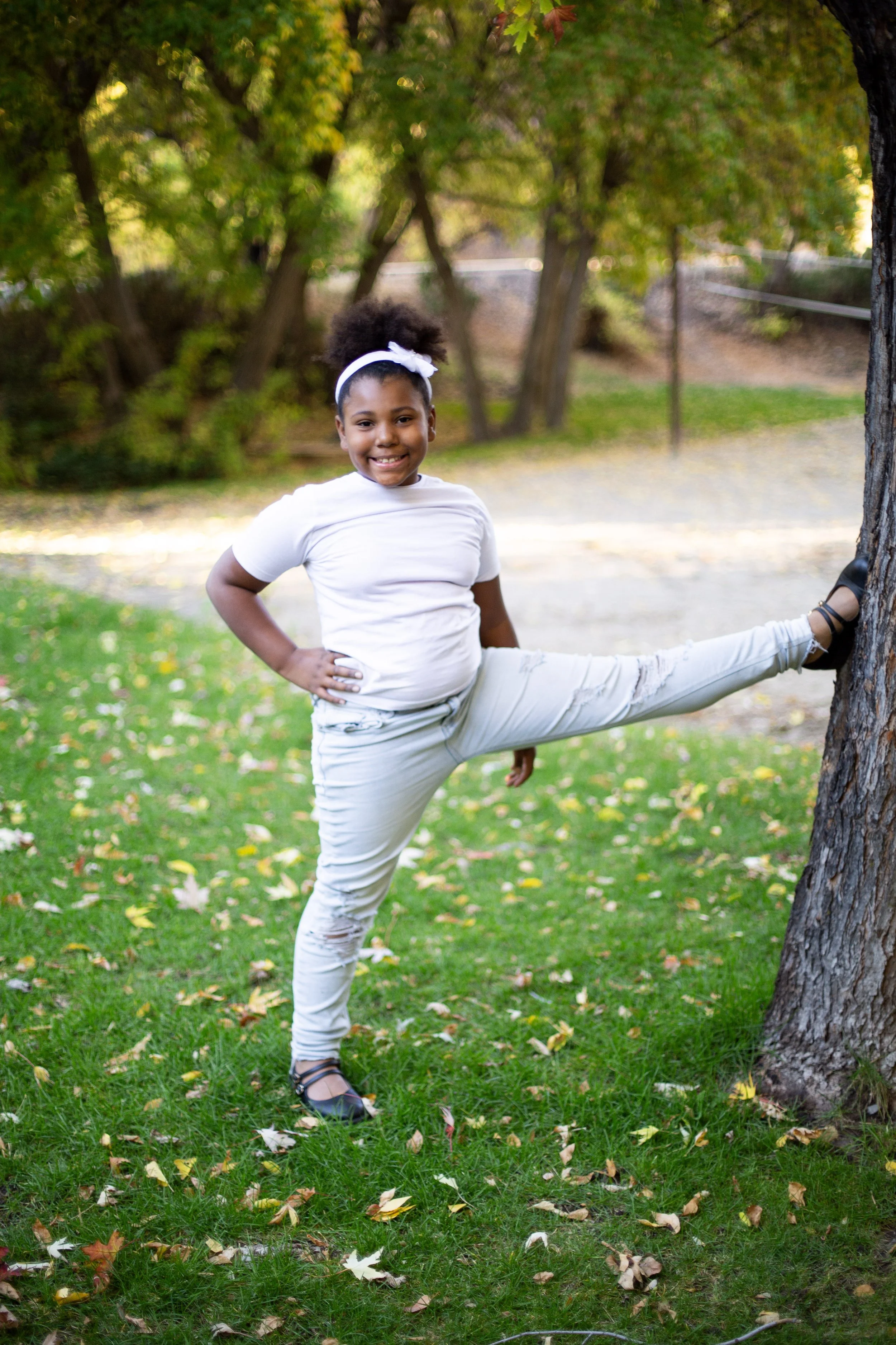 A young girl with curly hair in a white headband, wearing a white shirt and distressed white jeans, stands outdoors with one hand on her hip and the other leg stretched out against a tree, smiling at the camera in a park with green grass and trees.