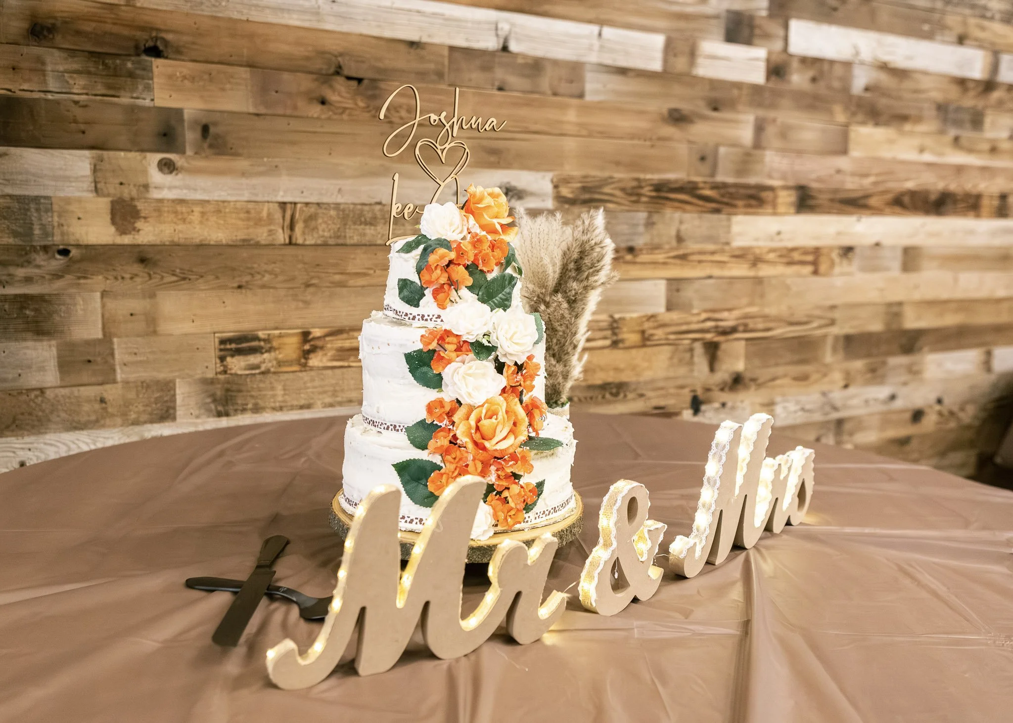 A wedding cake decorated with white frosting, orange and white flowers, and green leaves. Behind the cake is a wooden wall, and the cake is placed on a gold-colored base. There is a decorative sign in front of the cake that reads "love" with a heart 