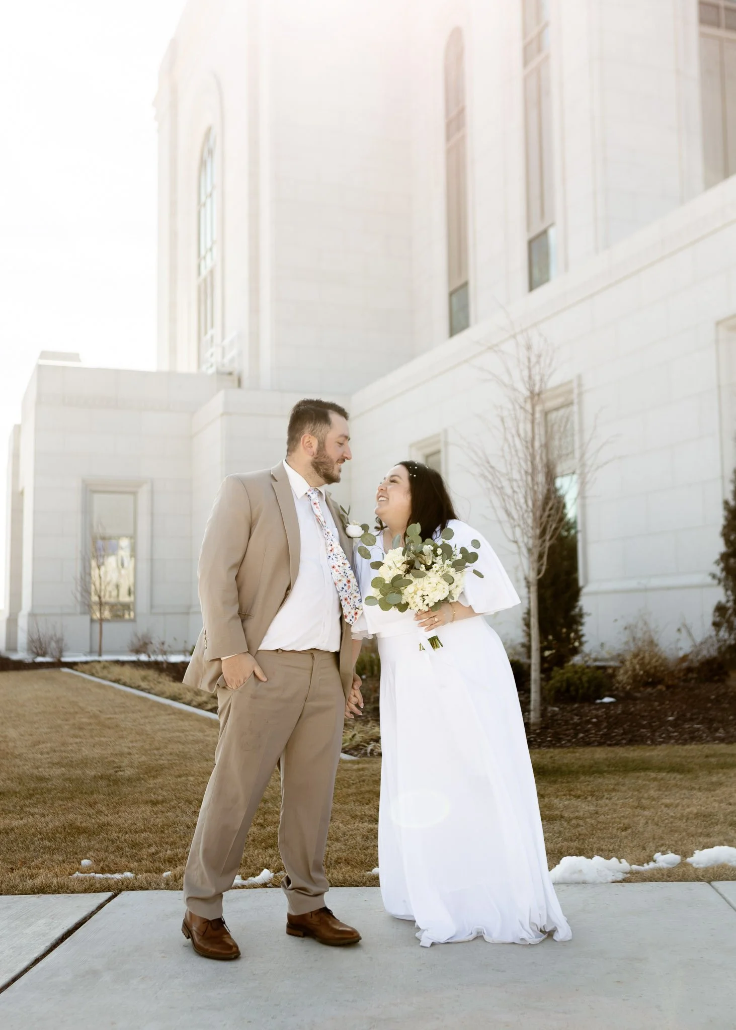 A couple dressed in wedding attire standing outdoors on a sidewalk, looking at each other and smiling, with a modern white building in the background and sunlight shining from the left.