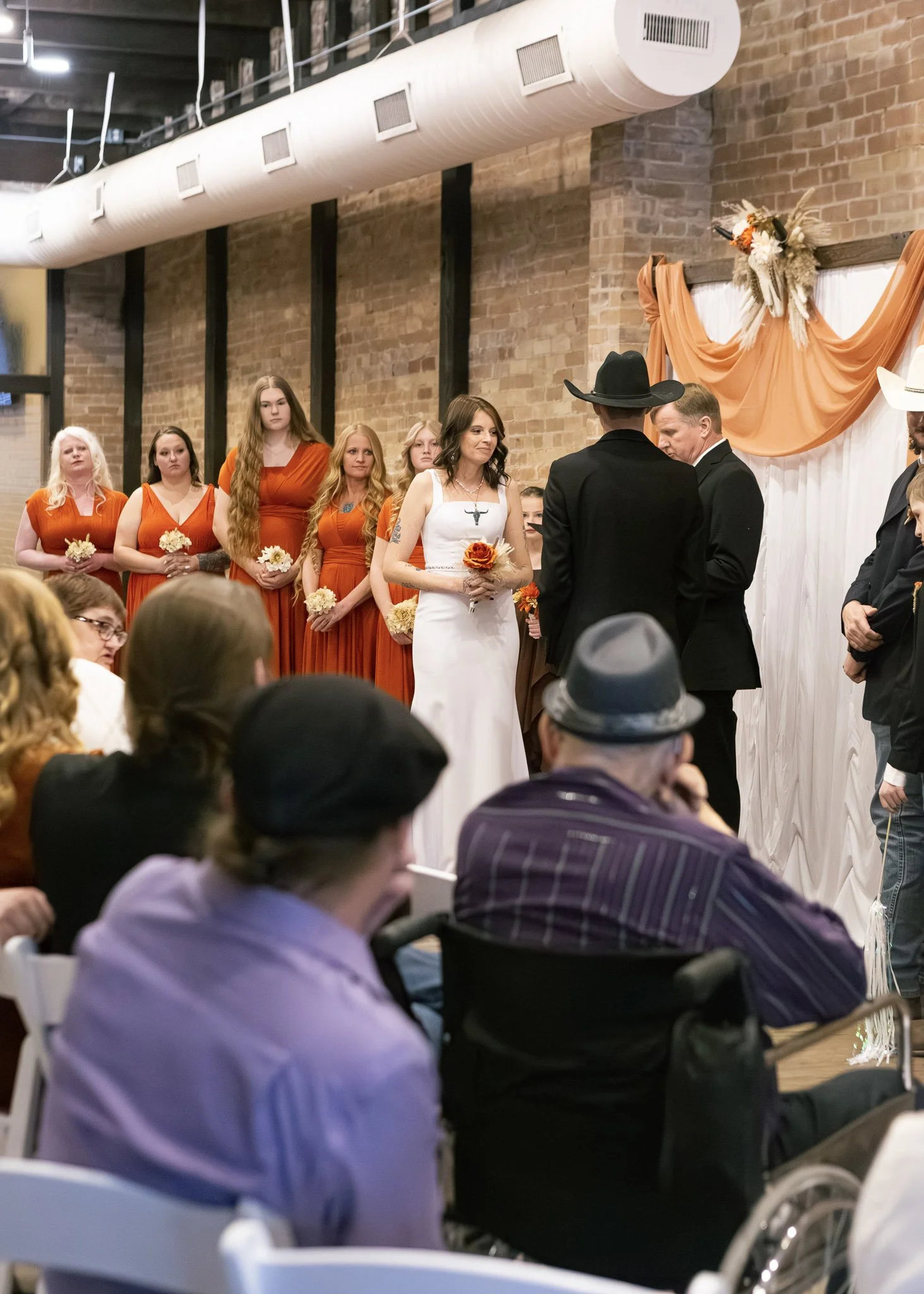 A wedding ceremony with guests seated, including a woman in a white dress holding a flower and an altar decorated with orange fabric and flowers.