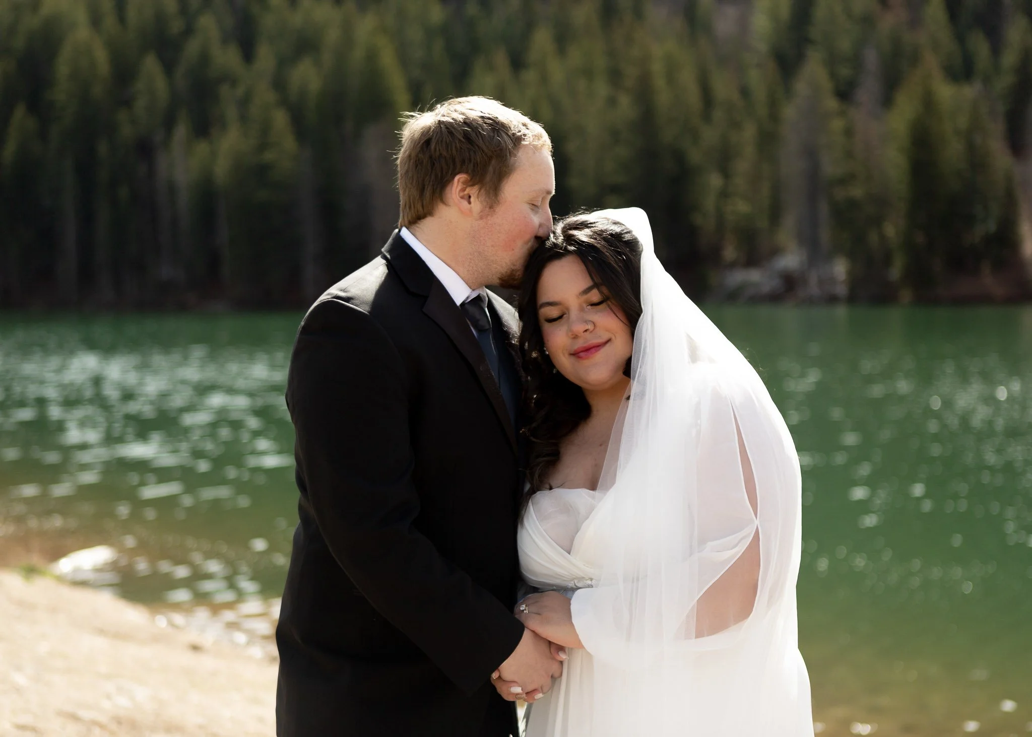 A couple dressed in wedding attire embracing by a lake with a forested background.