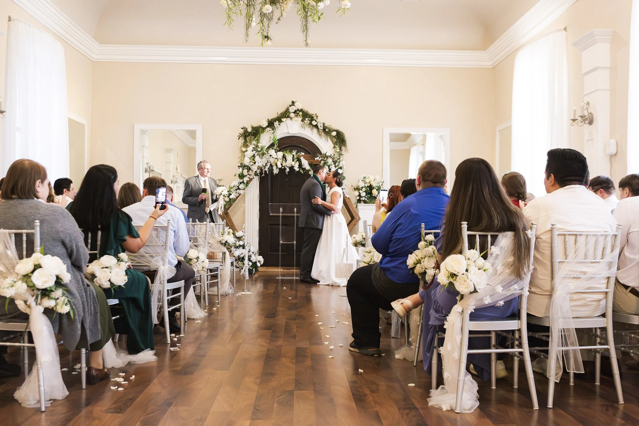 A couple at their wedding ceremony kissing in front of an arch decorated with white flowers and greenery, with guests seated on either side watching and taking photos.