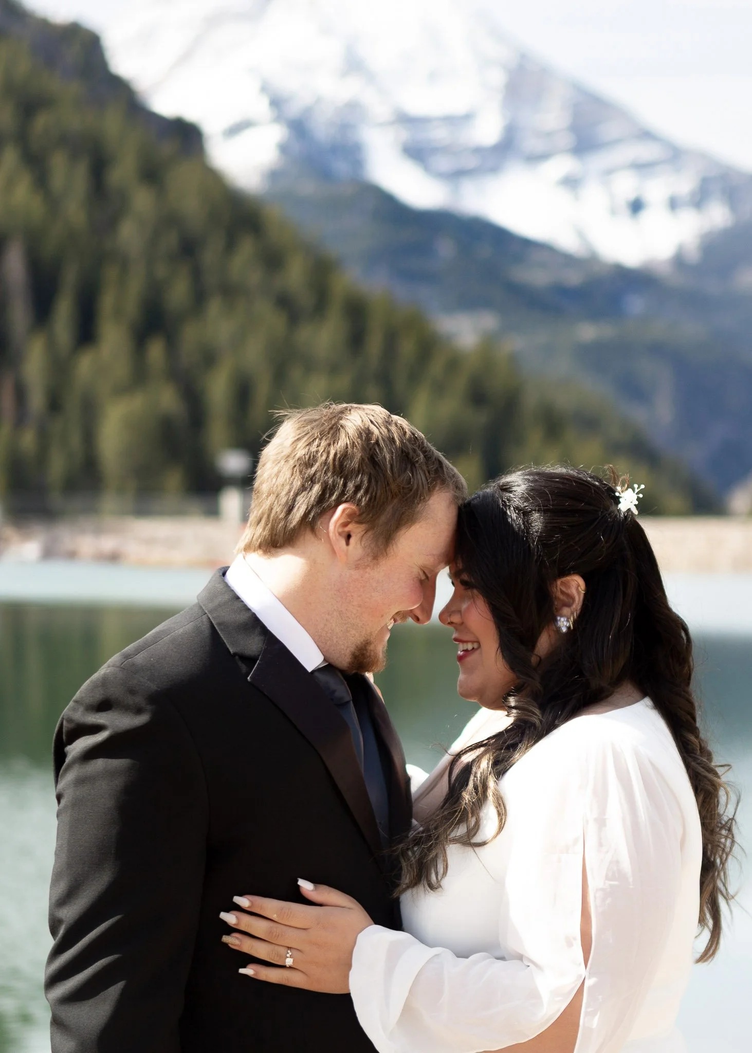 A newly married couple stands outdoors by a lake, embracing and touching foreheads, with mountains and greenery in the background.