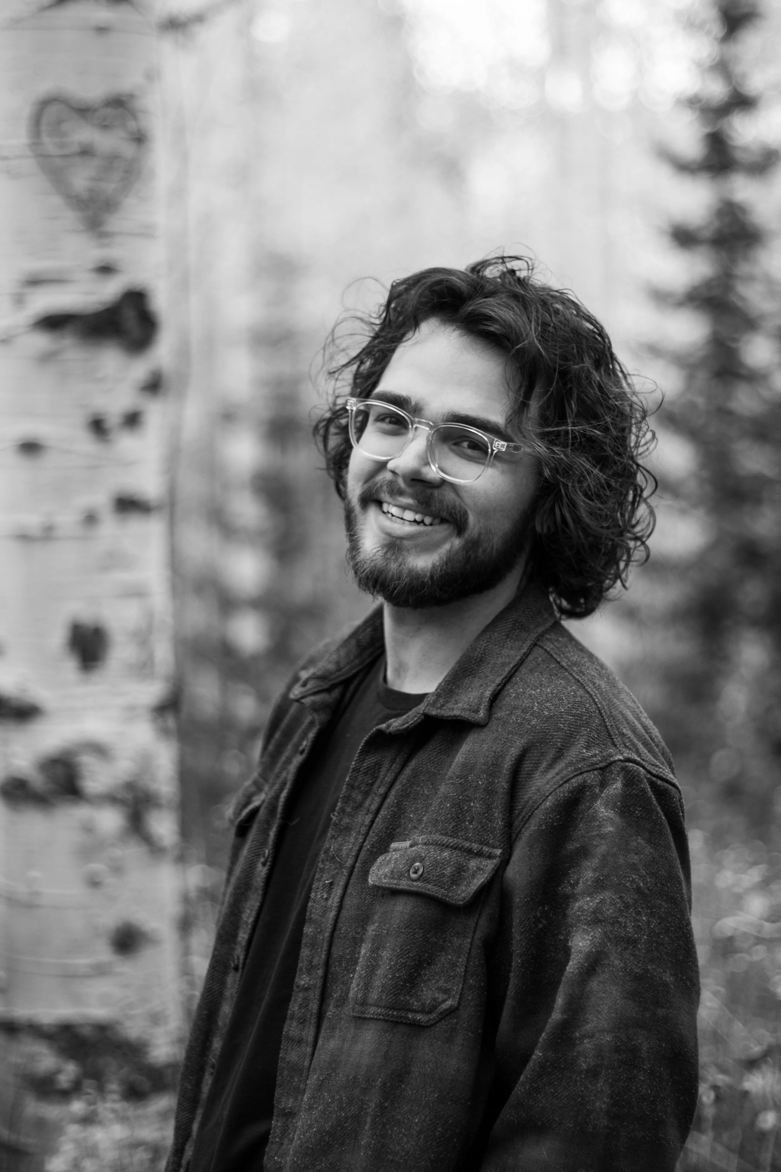 A smiling man with long curly hair, beard, and glasses, standing outdoors in front of a blurred forest background.