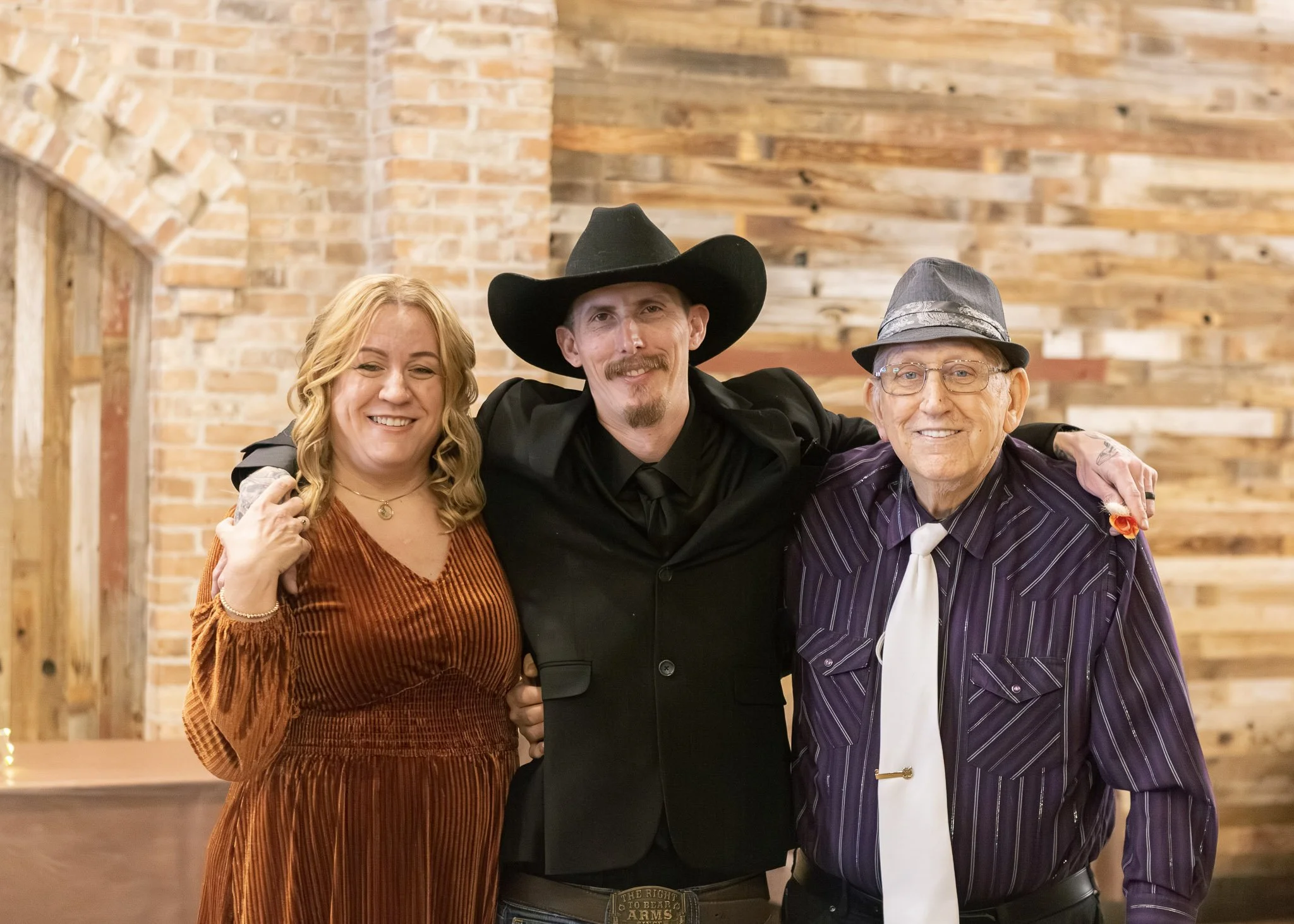 Three people smiling and posing for a photo in front of a brick and wood wall, with two women on each side of a man wearing a black cowboy hat and a black blazer.