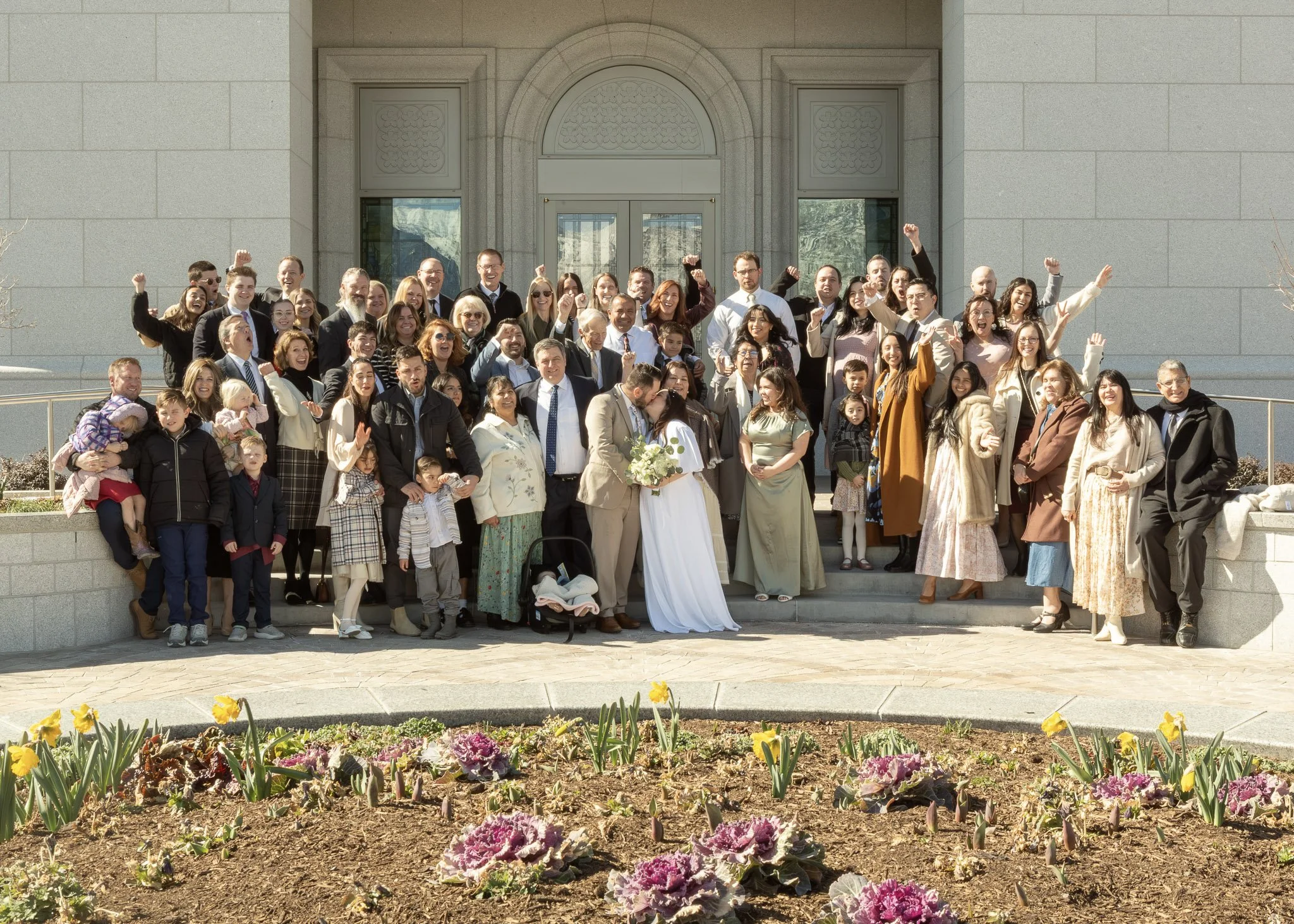 A large group of people, including men, women, and children, celebrating outside a building with stone walls and an arched entrance. Some people are raising their fists or arms, and a bride and groom are in the front center holding flowers.
