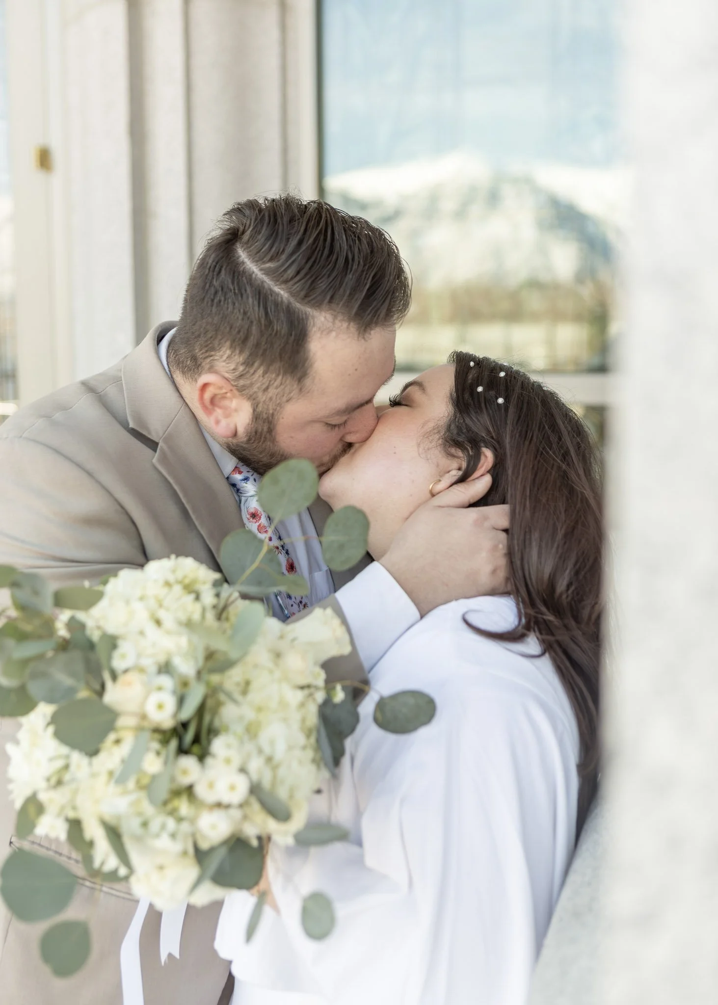 A couple kissing at a wedding, with a bouquet of white flowers and greenery in the foreground.