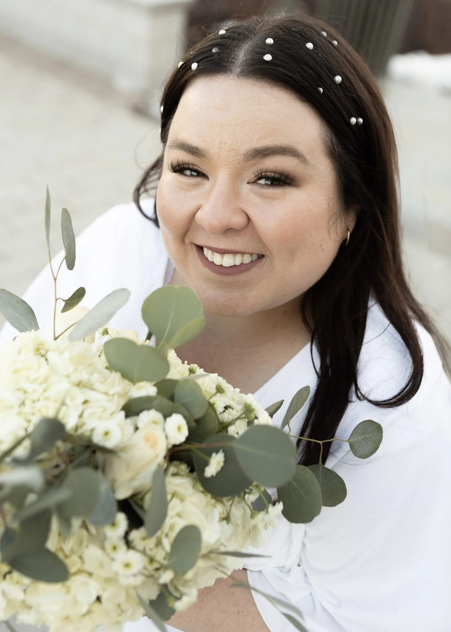 A woman with dark hair decorated with white pearls, smiling while holding a bouquet of white flowers and eucalyptus leaves.