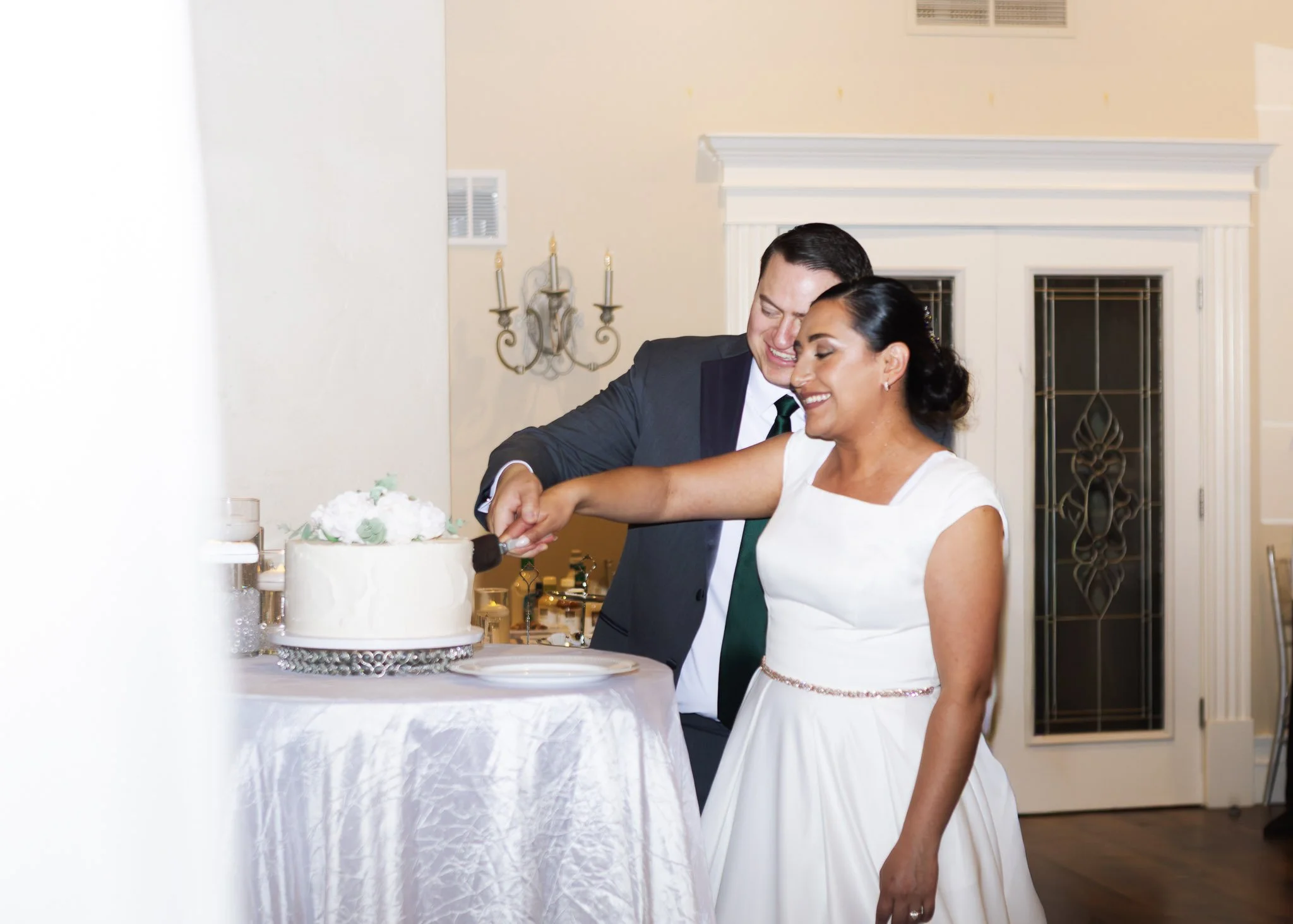 A newlywed couple cutting their wedding cake together during their wedding reception. The groom is dressed in a dark suit and tie, and the bride is wearing a white wedding dress. They are smiling and appear happy.