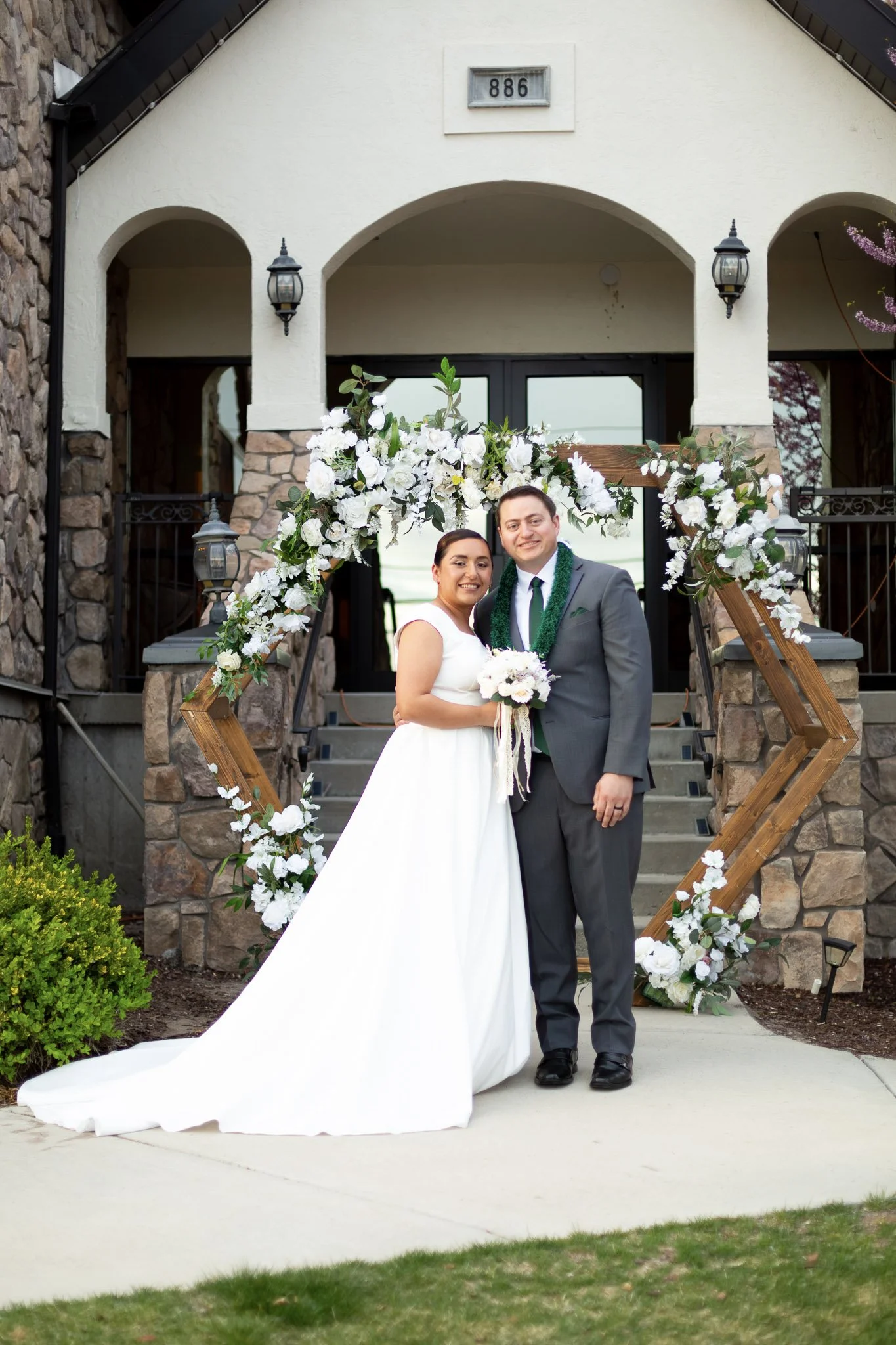 A bride and groom standing outside a building with stone and stucco walls, decorated with a floral archway. The bride is in a white wedding dress, holding a bouquet, and the groom is in a gray suit with a green garland around his neck, smiling.
