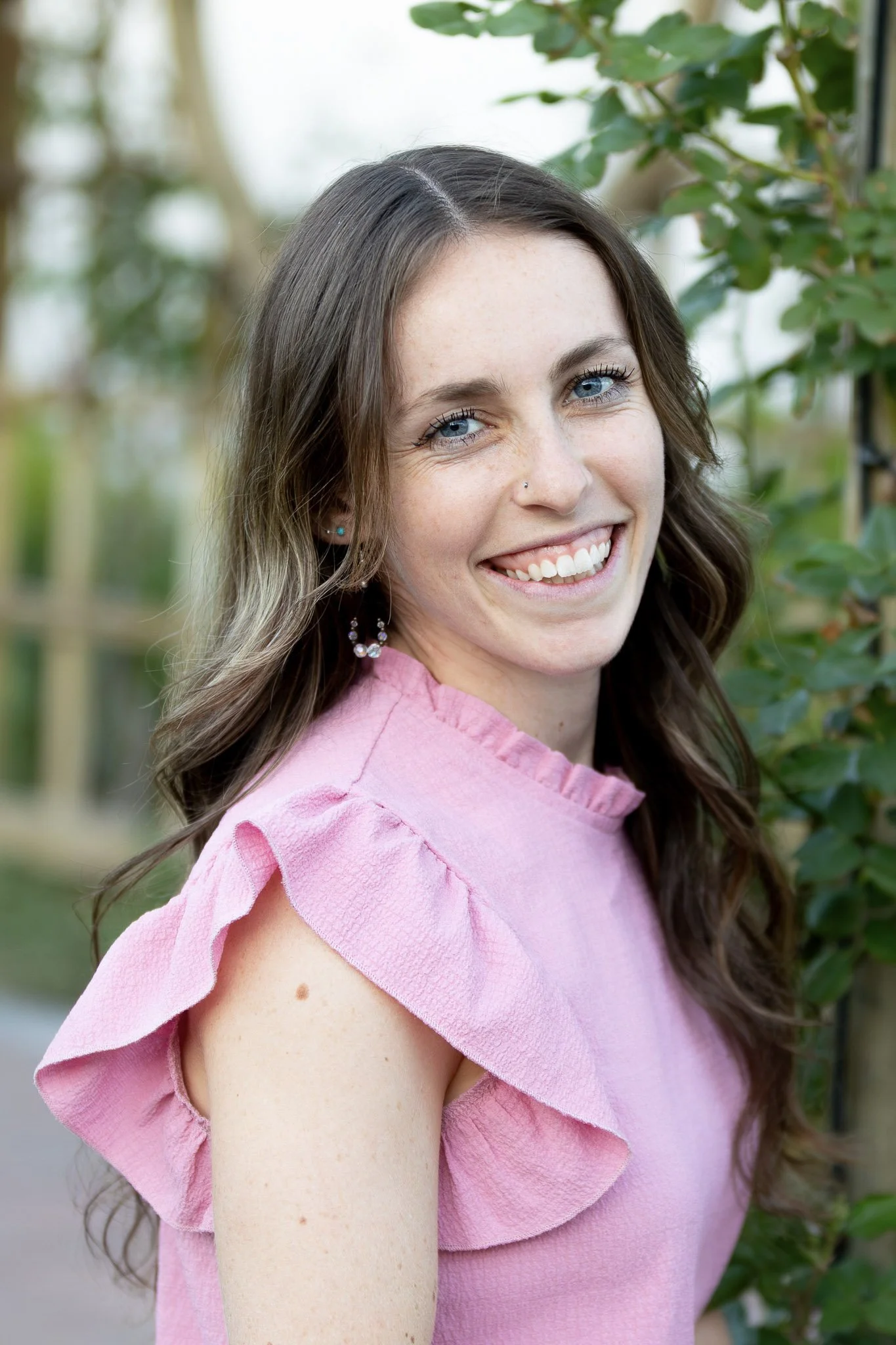 A smiling woman with long, wavy brown hair wearing a pink ruffled sleeve top standing outdoors near green foliage.