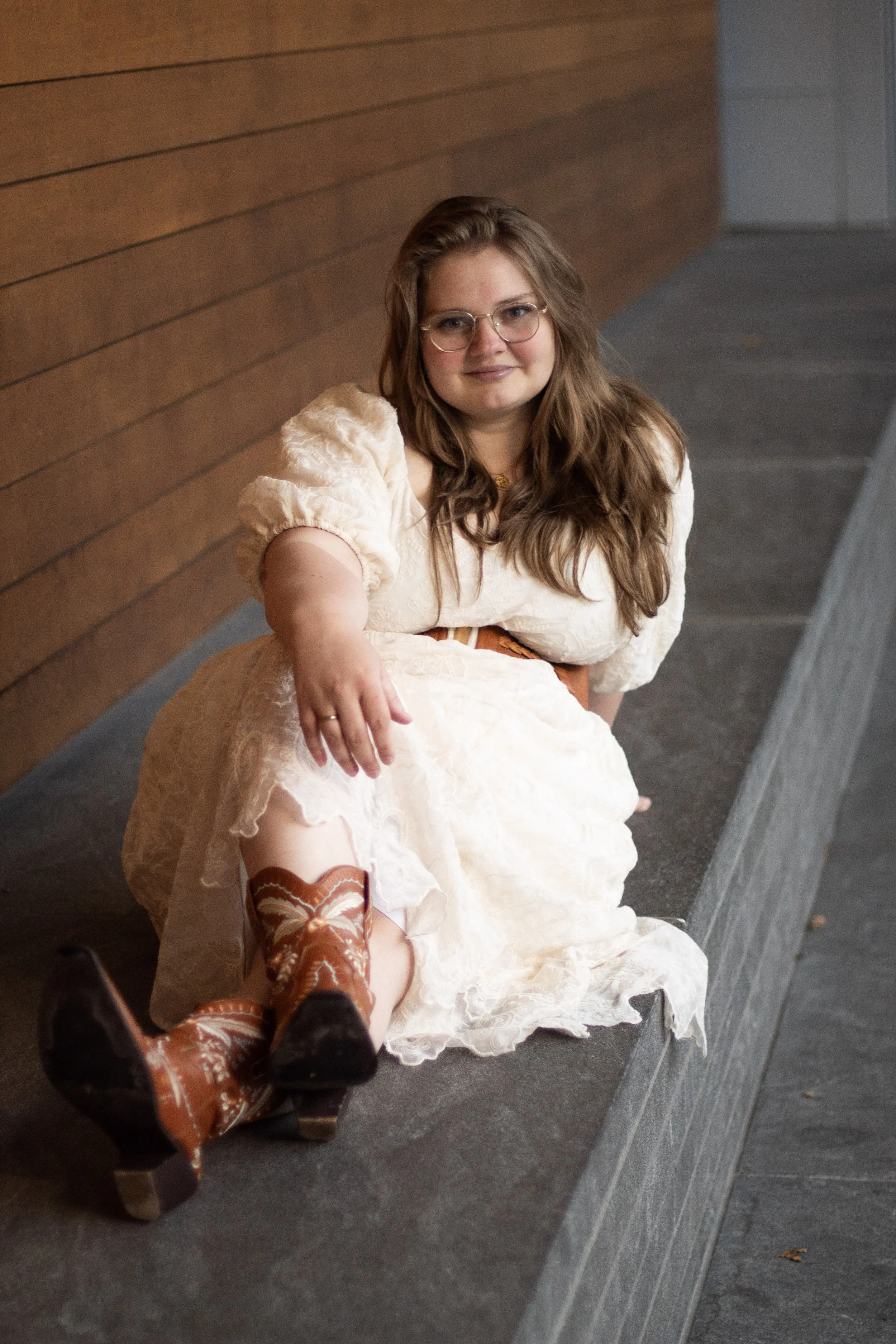 A woman with long, wavy brown hair and glasses sitting on a low wall, wearing a cream-colored dress and cowboy boots, outdoors near a wooden wall.