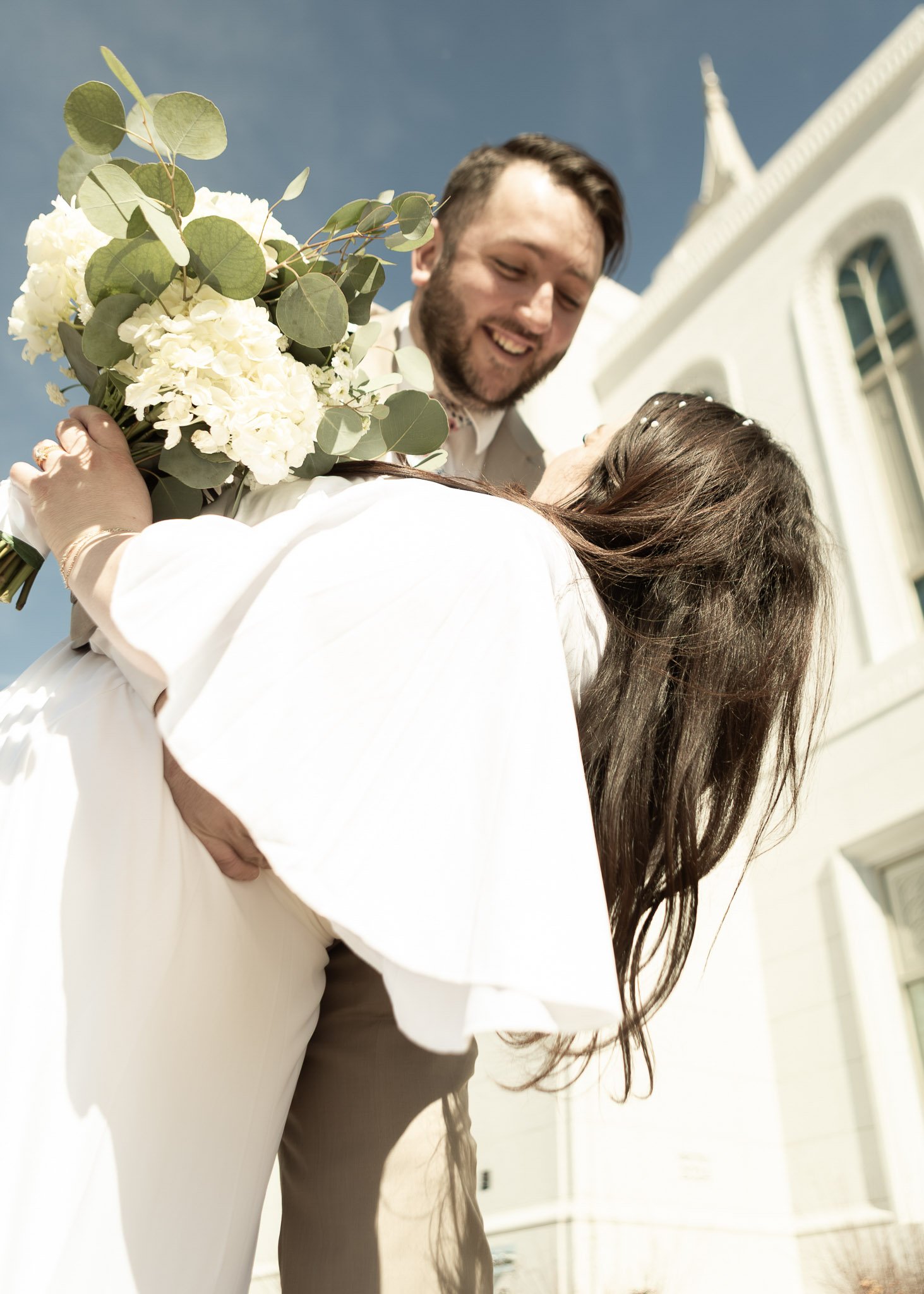 A man in formal attire embracing a woman holding a bouquet of white flowers, outside a white building with a clear blue sky.