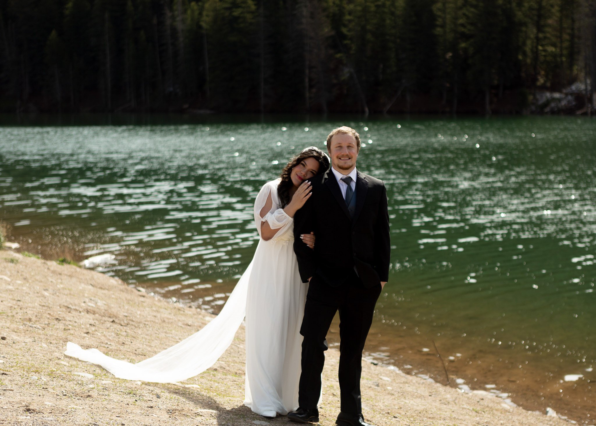 A newlywed couple poses on a lake beach surrounded by forest, with the bride in a white dress and the groom in a black tuxedo, both smiling.