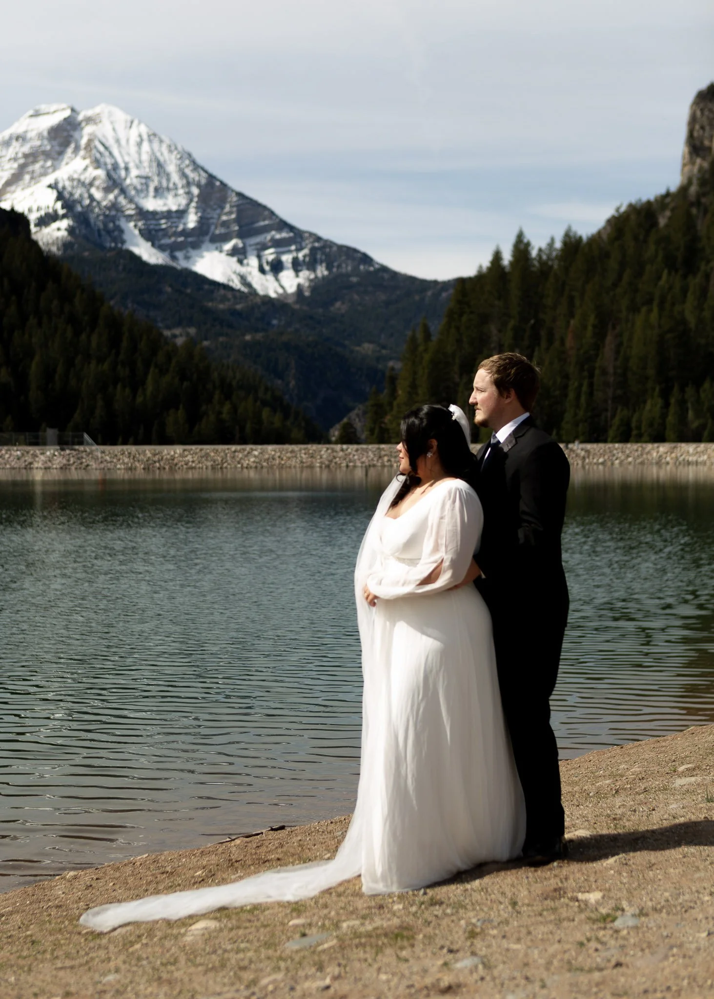 A bride and groom stand by a lake with mountains in the background. The bride wears a white dress and veil, and the groom is in a black tuxedo. The scene is outdoors with natural scenery.