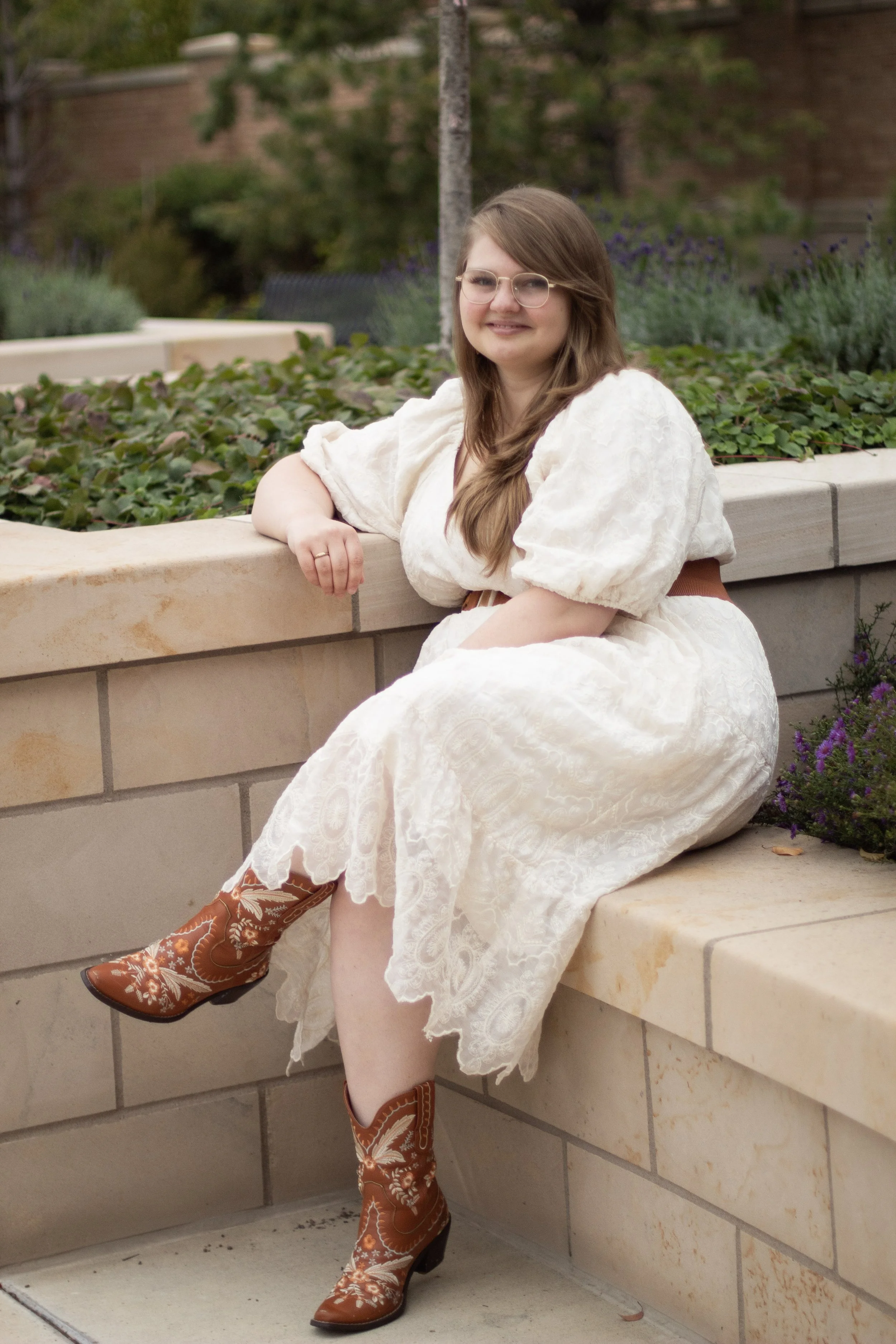 A woman sitting on a stone ledge in a garden, wearing a cream-colored lace dress and cowboy boots, smiling at the camera.