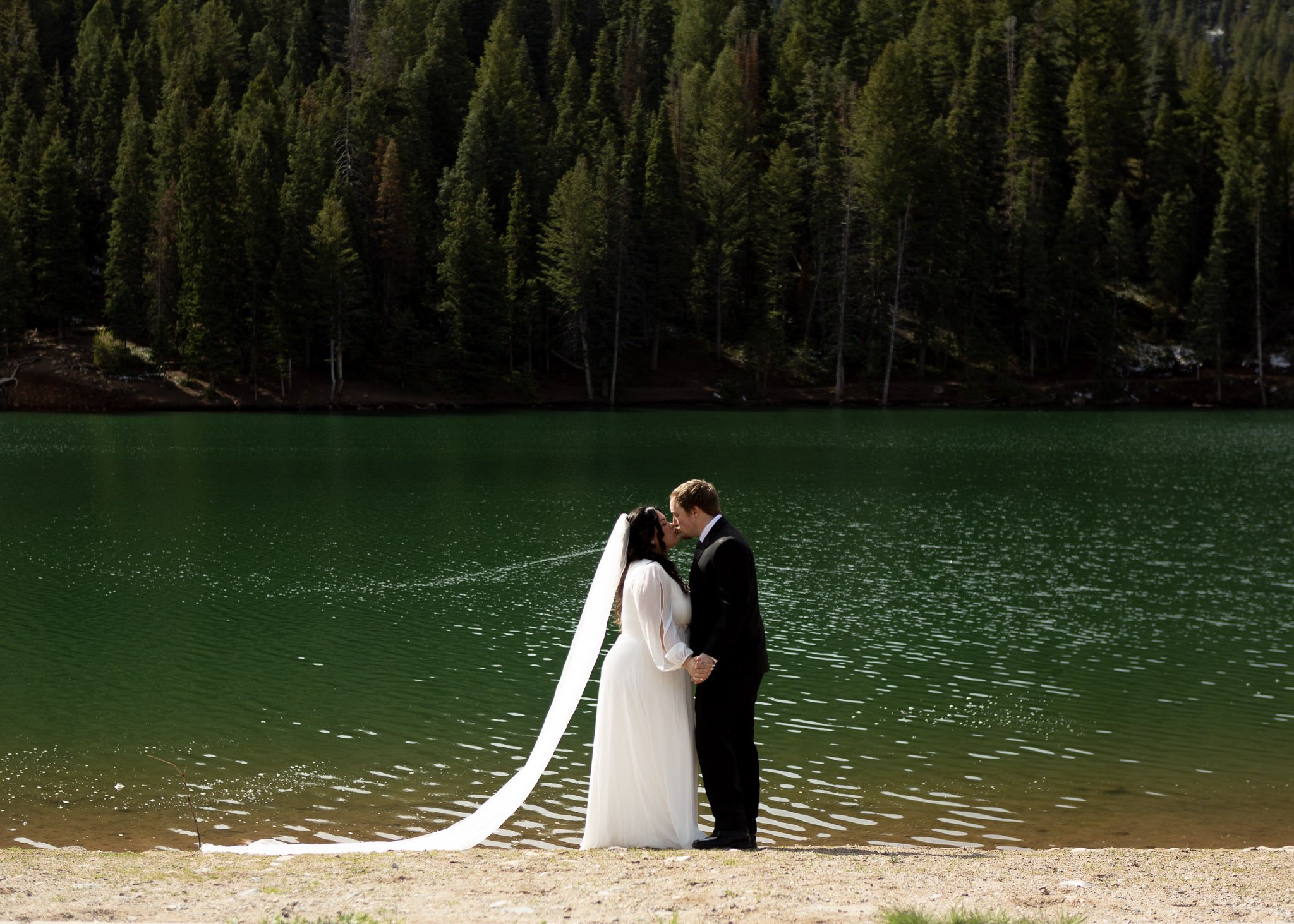 A bride and groom standing close together by a lake, about to kiss, with trees in the background.