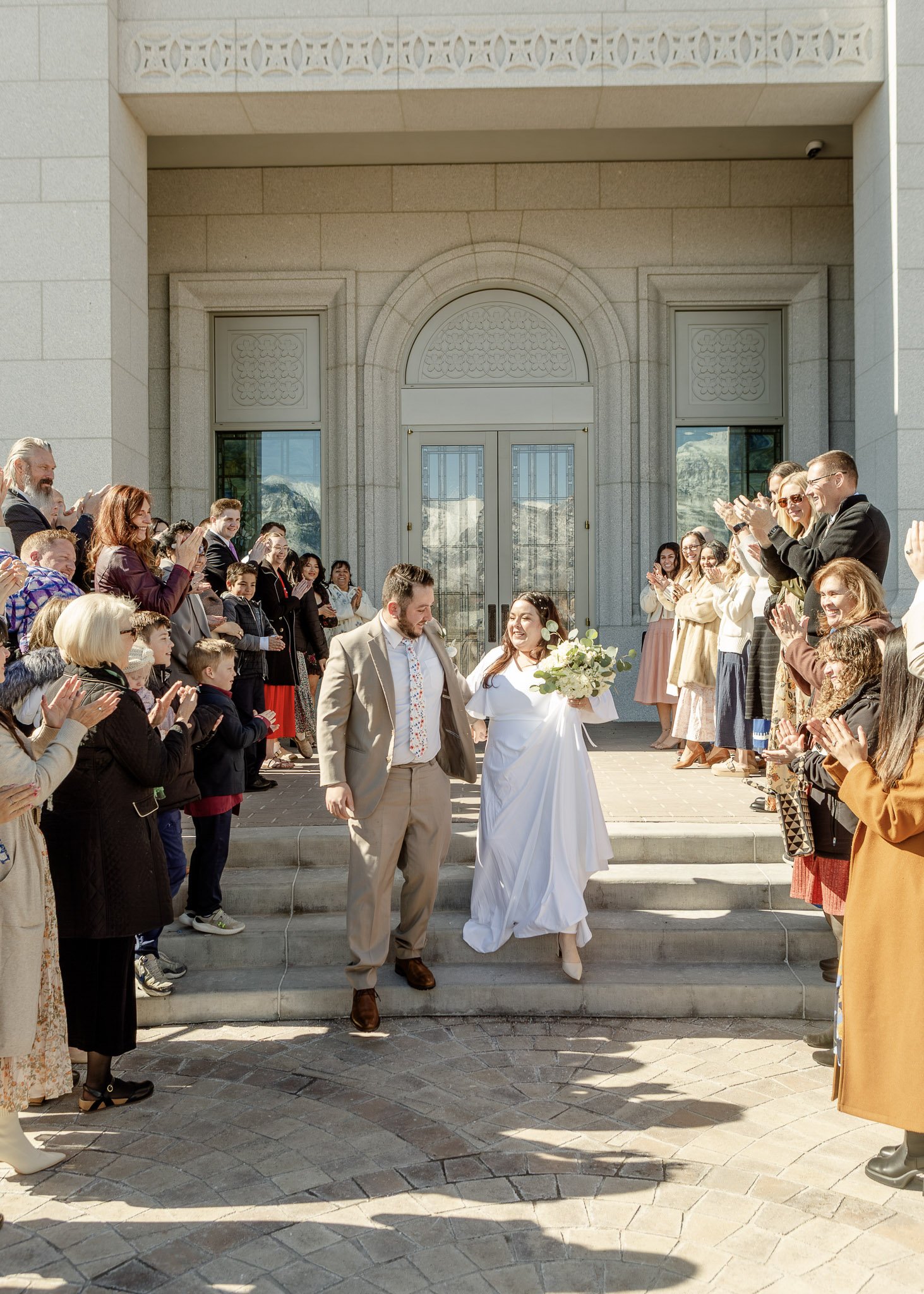 A newly married couple is walking down the stairs outside a building while guests celebrate and applaud. The bride is wearing a white wedding dress and holding a bouquet, and the groom is dressed in a beige suit. The guests are standing on either sid