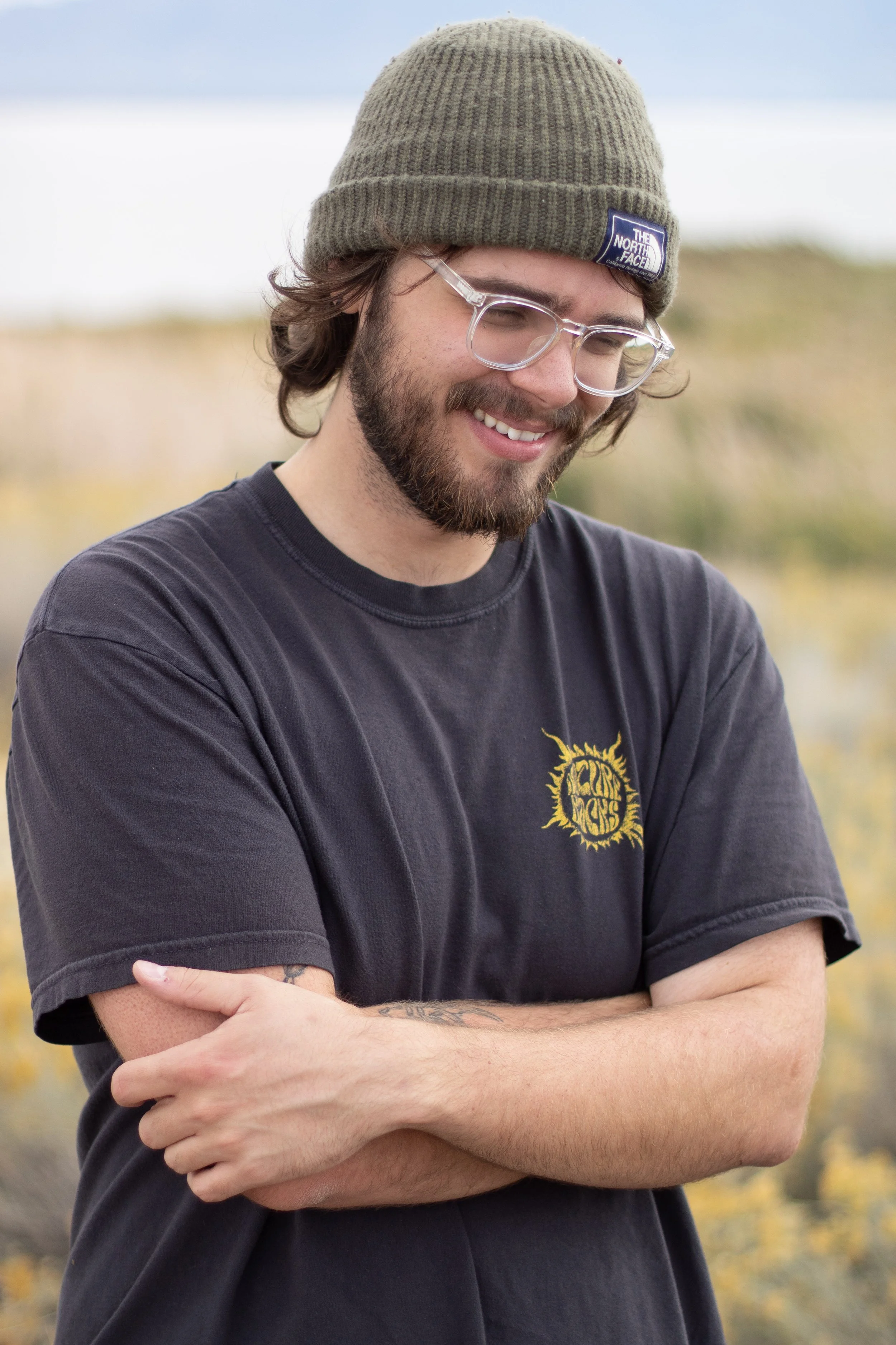 A smiling man wearing a green knit beanie, clear glasses, and a black T-shirt with yellow graphic design, standing outdoors with a blurred background of yellow and brown plants.