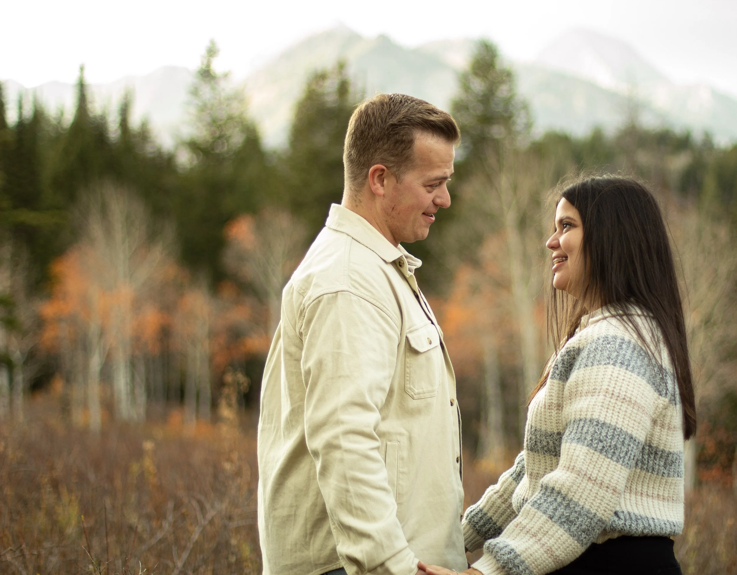 A couple holding hands and smiling at each other outdoors during fall in front of fall foliage with mountains in the background.