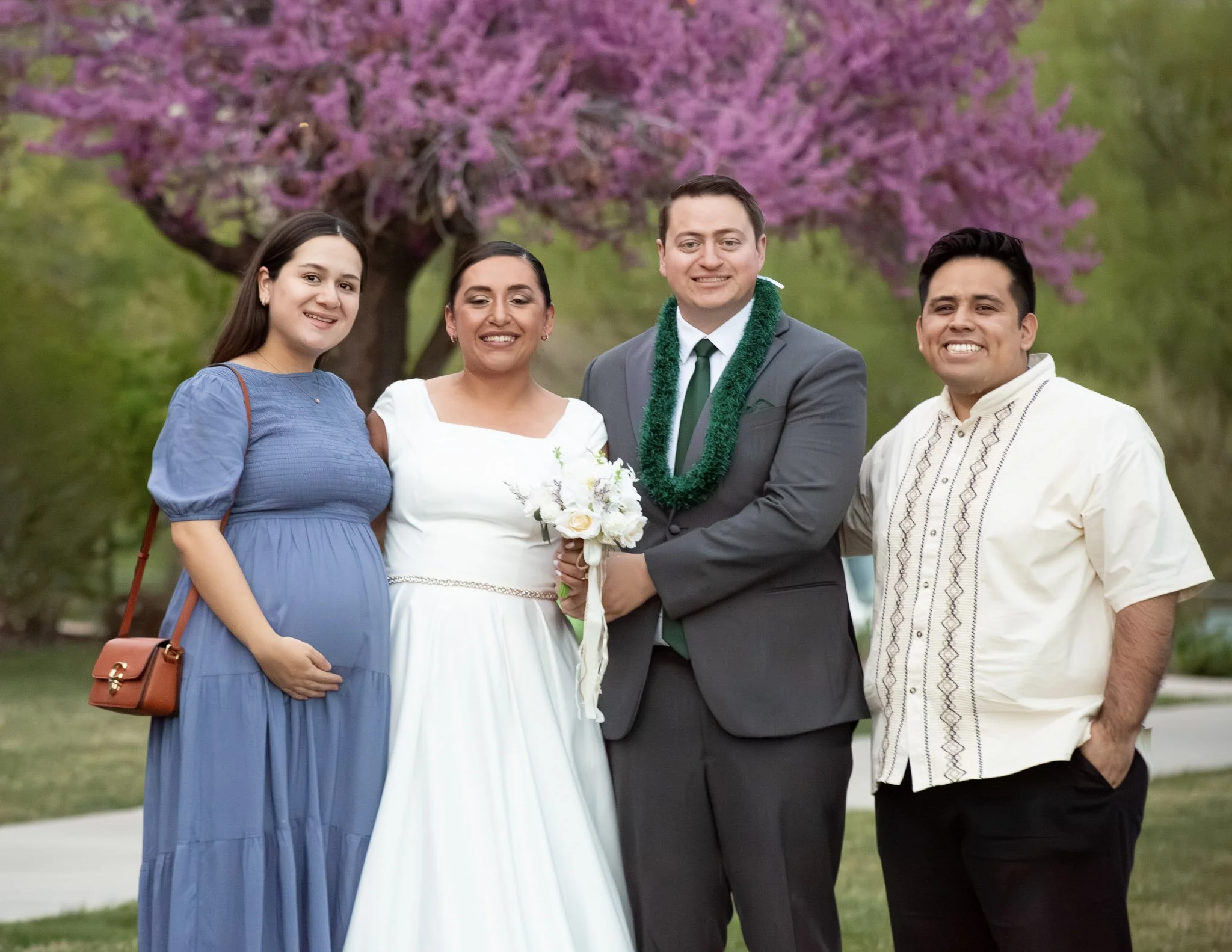 A group of four people standing outdoors near a pink flowering tree. Two women and two men, smiling, dressed in semi-formal or casual attire, with a woman holding a bouquet. The man in the middle wears a suit with a green tie and a green garland, sug