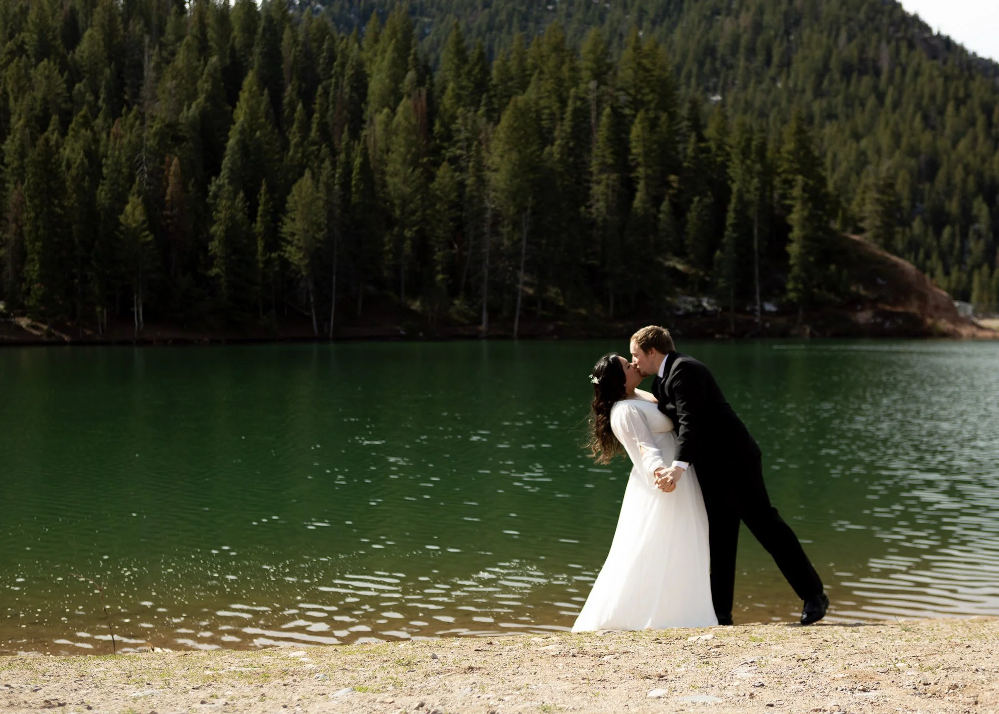 A bride and groom sharing a kiss by a green lake with a dense forested mountain in the background.