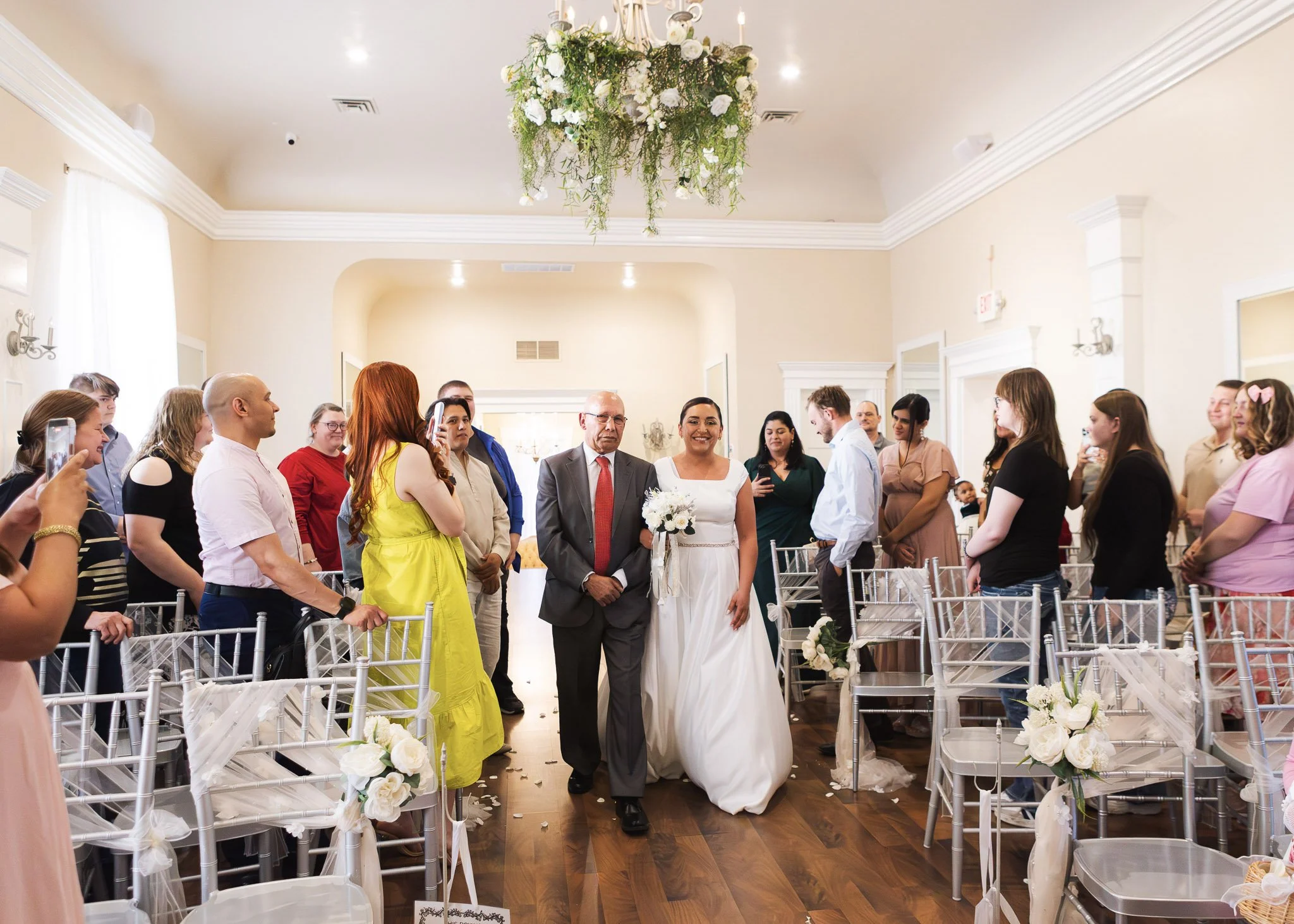A bride in a white wedding dress and a groom in a suit walk down the aisle in a decorated wedding venue with guests on both sides, floral arrangements, and a chandelier hanging from the ceiling.